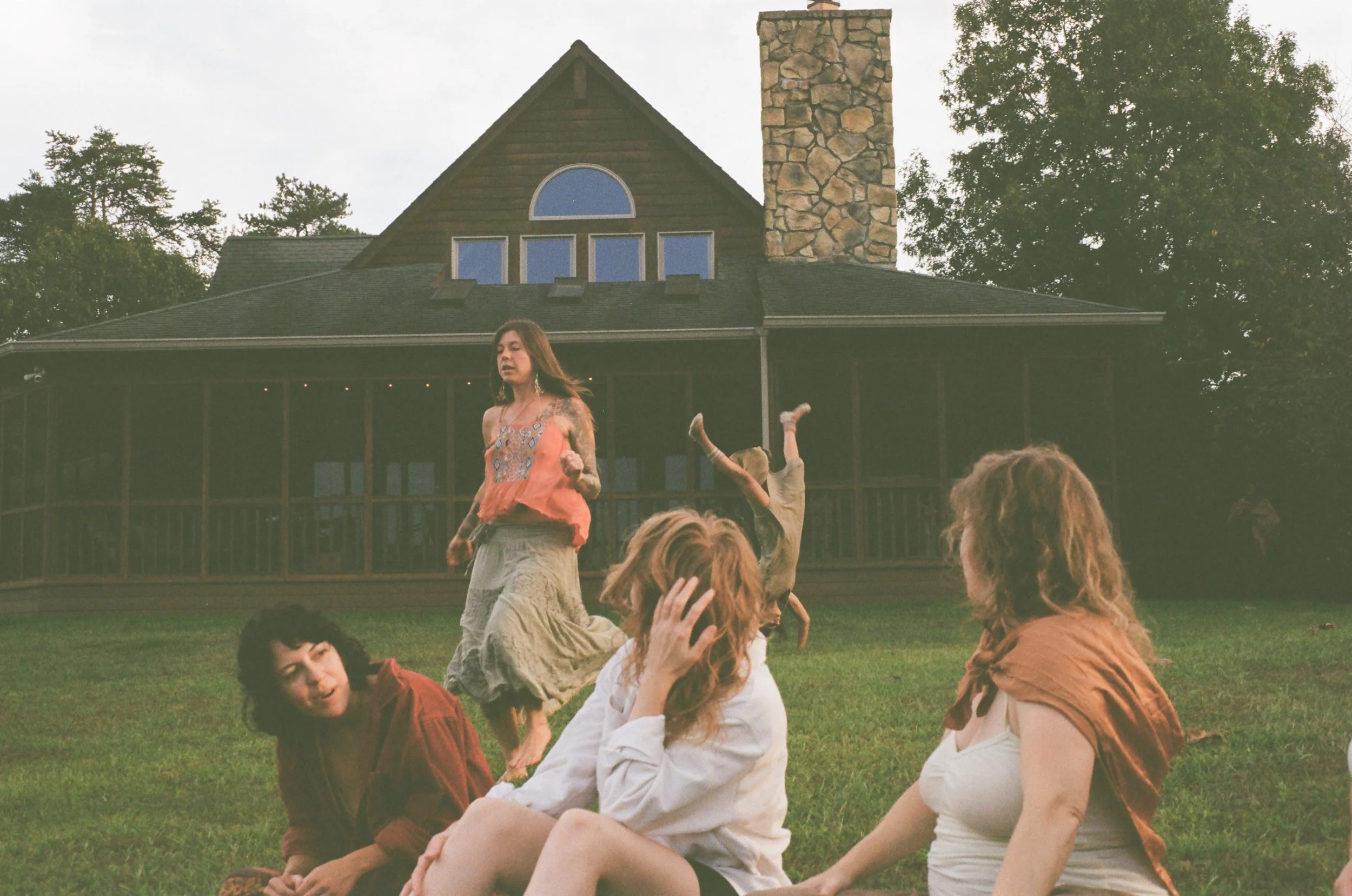 A group of women sitting and standing on a grassy lawn in front of a large house with a chimney, large windows, and a screened porch, during sunset or late afternoon.