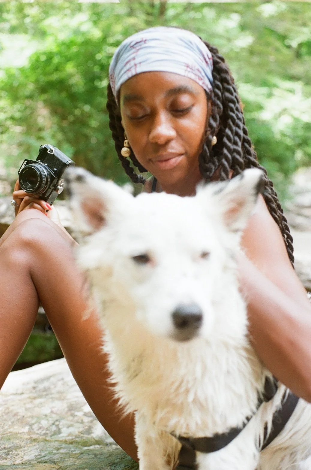 A woman with braided hair and earrings sitting outdoors with a white dog. She is holding a camera and looking down at the dog.