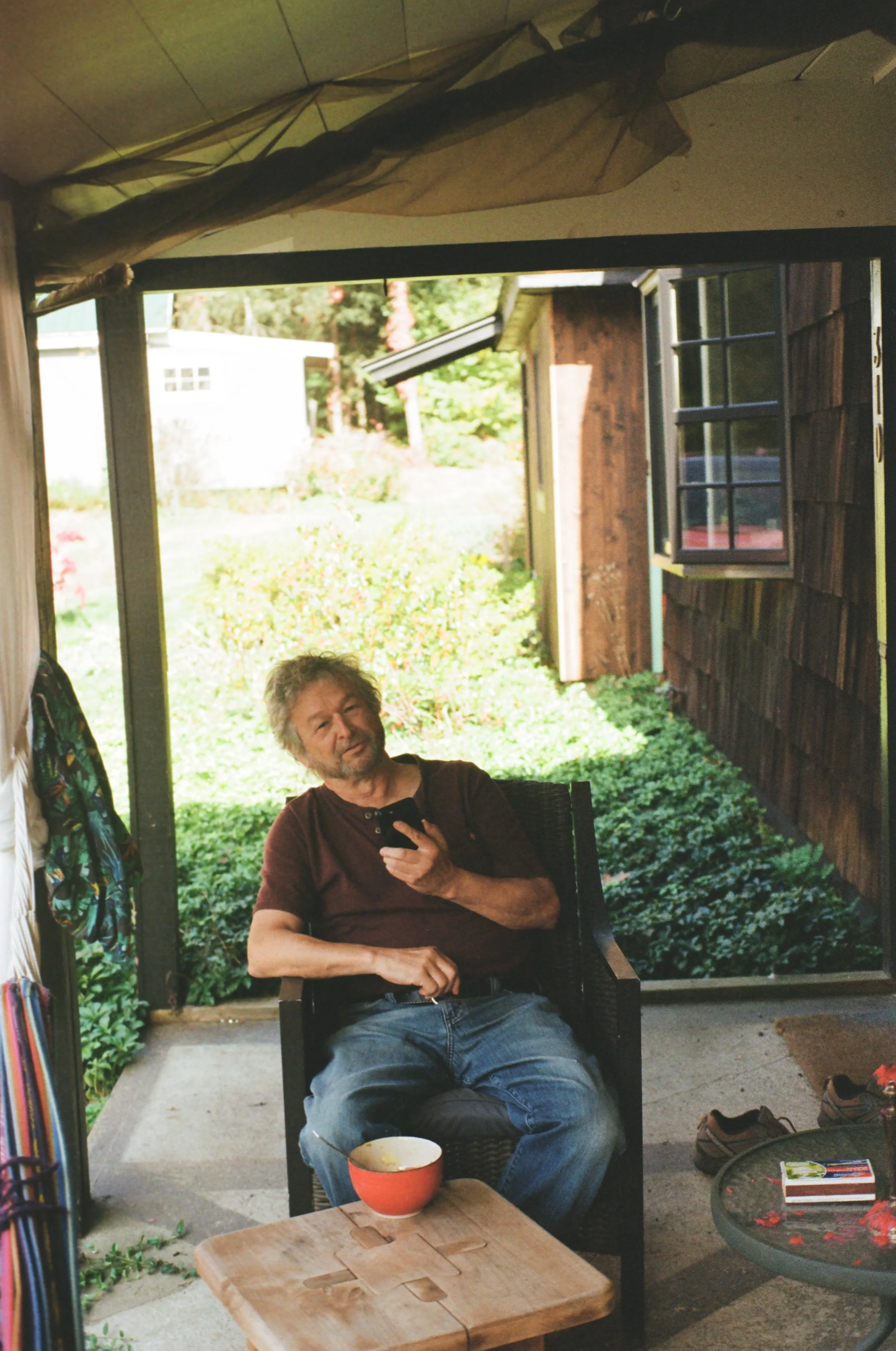 A man sitting outside on a patio, holding a phone, with shoes and a bowl on the table in front of him.