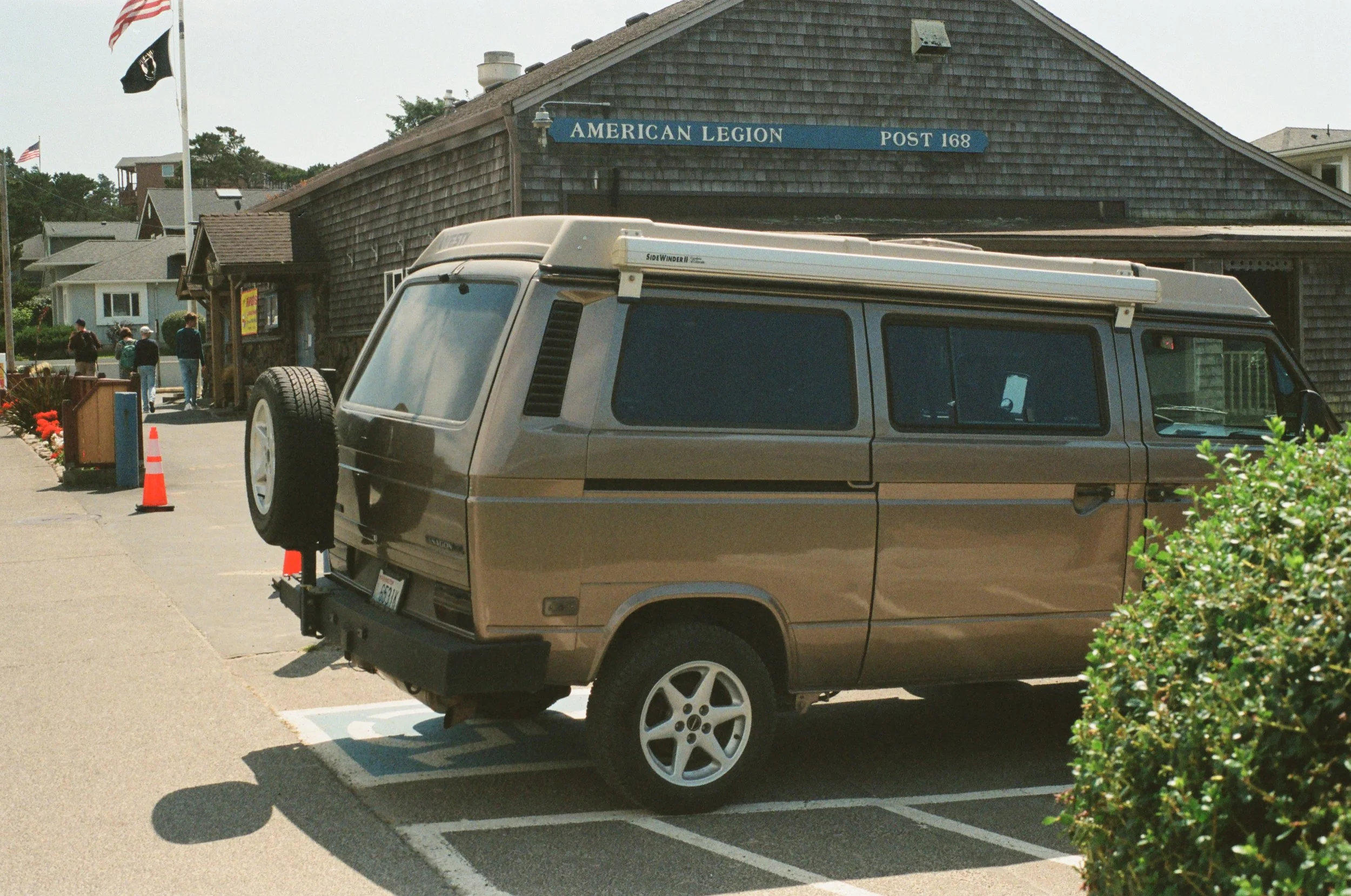 A vintage brown van parked in a parking lot in front of an American Legion building. The van has a spare tire mounted on the back and a white roof. The parking lot has orange traffic cones and a small bush on the right side.