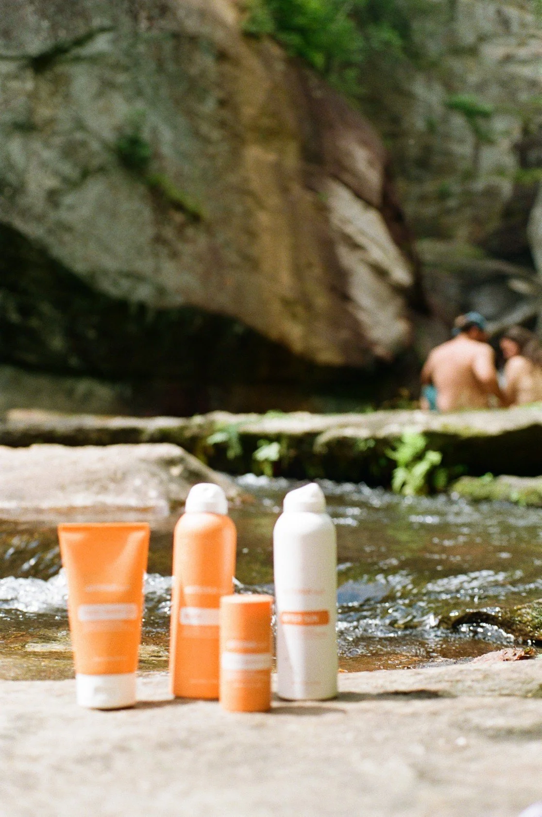 Arranged skincare products on a rock by a stream in a wooded area, with people in swimsuits in the background.