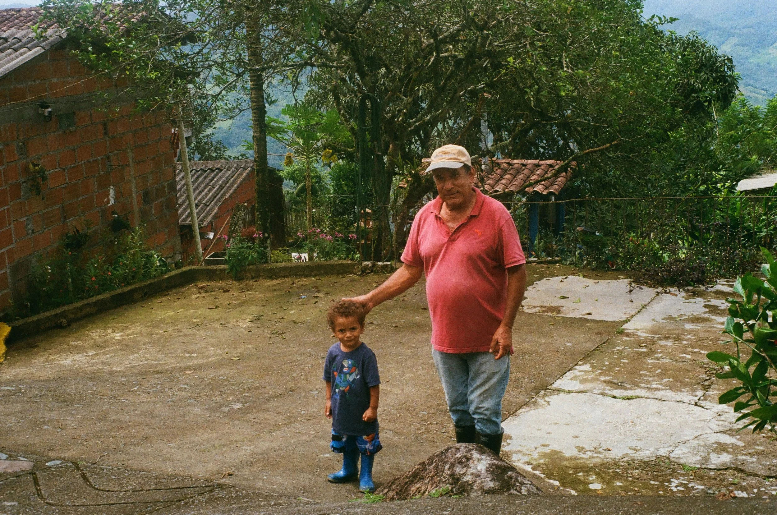 An elderly man with gray hair, wearing a pink polo shirt, blue jeans, and black boots, stands outdoors on a concrete surface, gently holding the hand of a young boy with curly hair, wearing a dark blue T-shirt with a graphic print, night pants, and b