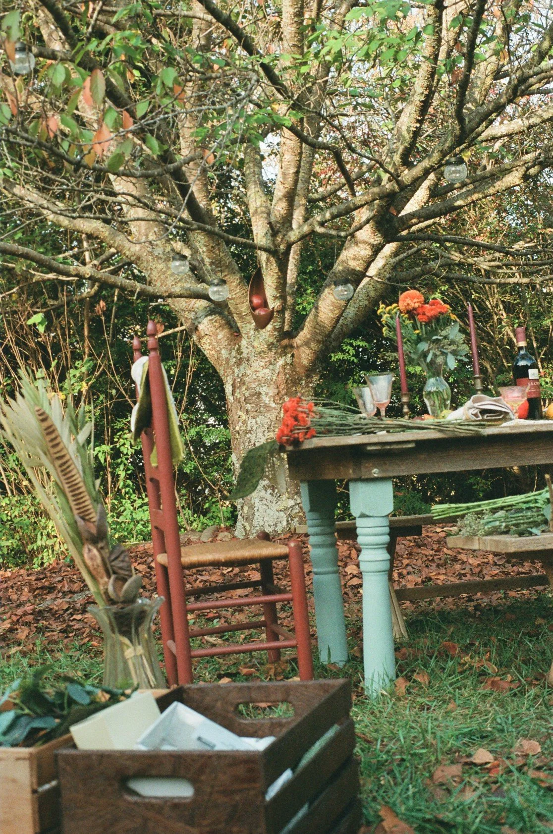 A garden setting with a large tree in the background and a rustic dining setup. The table has floral arrangements, candles, bottles, and glassware. A wooden chair with a white cushion and a small table with flowers are also visible.