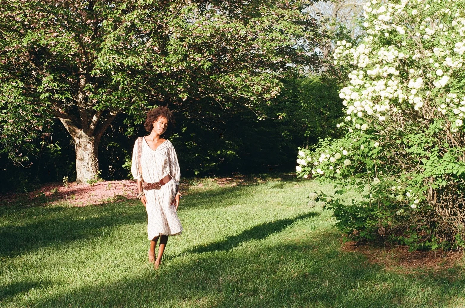 A woman stands on a grassy lawn under a tree with pink blossoms, wearing a light-colored dress and a brown belt, with greenery and blooming bushes in the background.