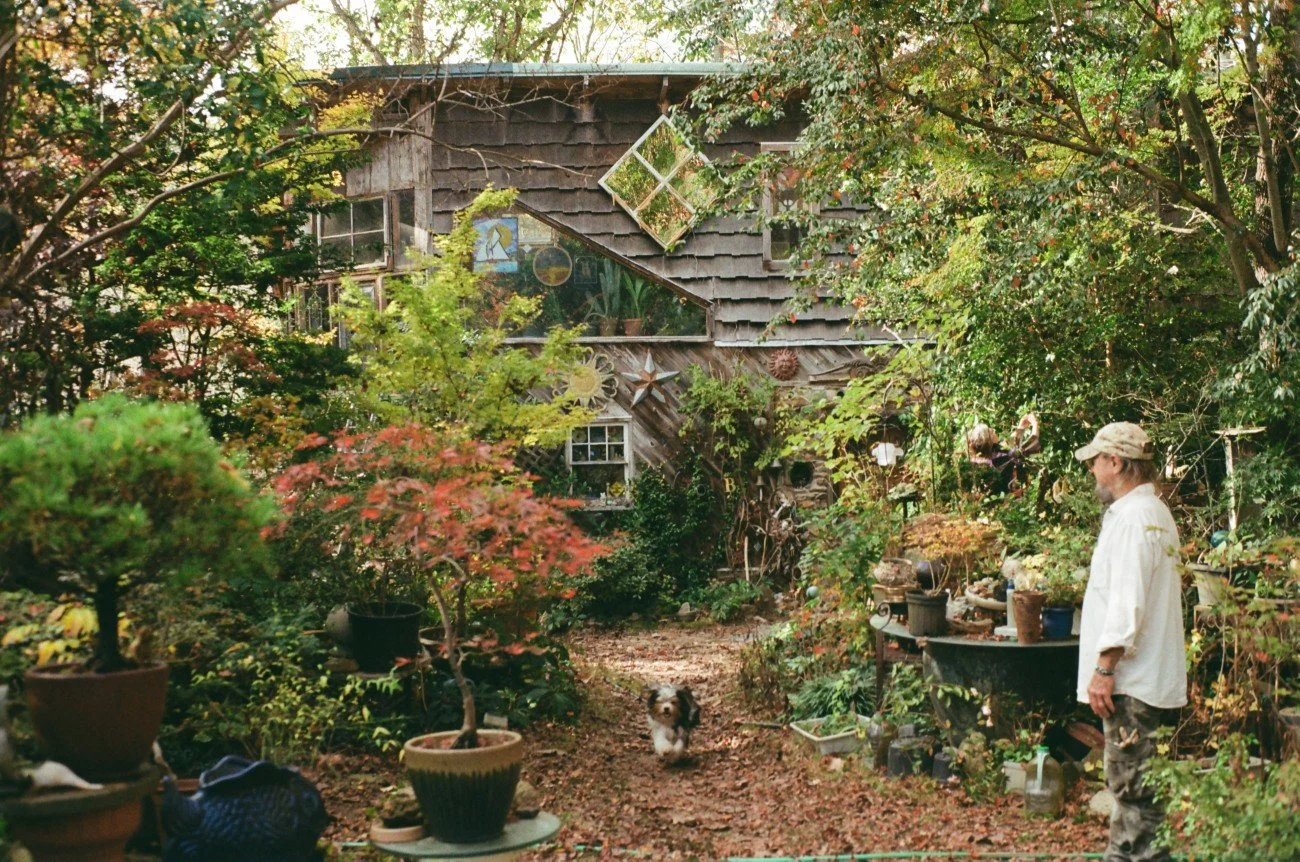 A man in a white shirt and camouflage pants is standing in a lush garden with potted plants and garden decorations, facing a small dog among the greenery, with a wooden house with decorative items on the exterior in the background.
