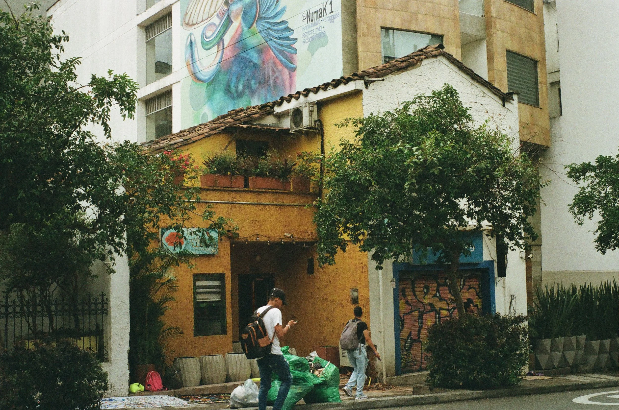 Street scene with two young men walking past a yellow building with a mural on the wall, trees, and street art.