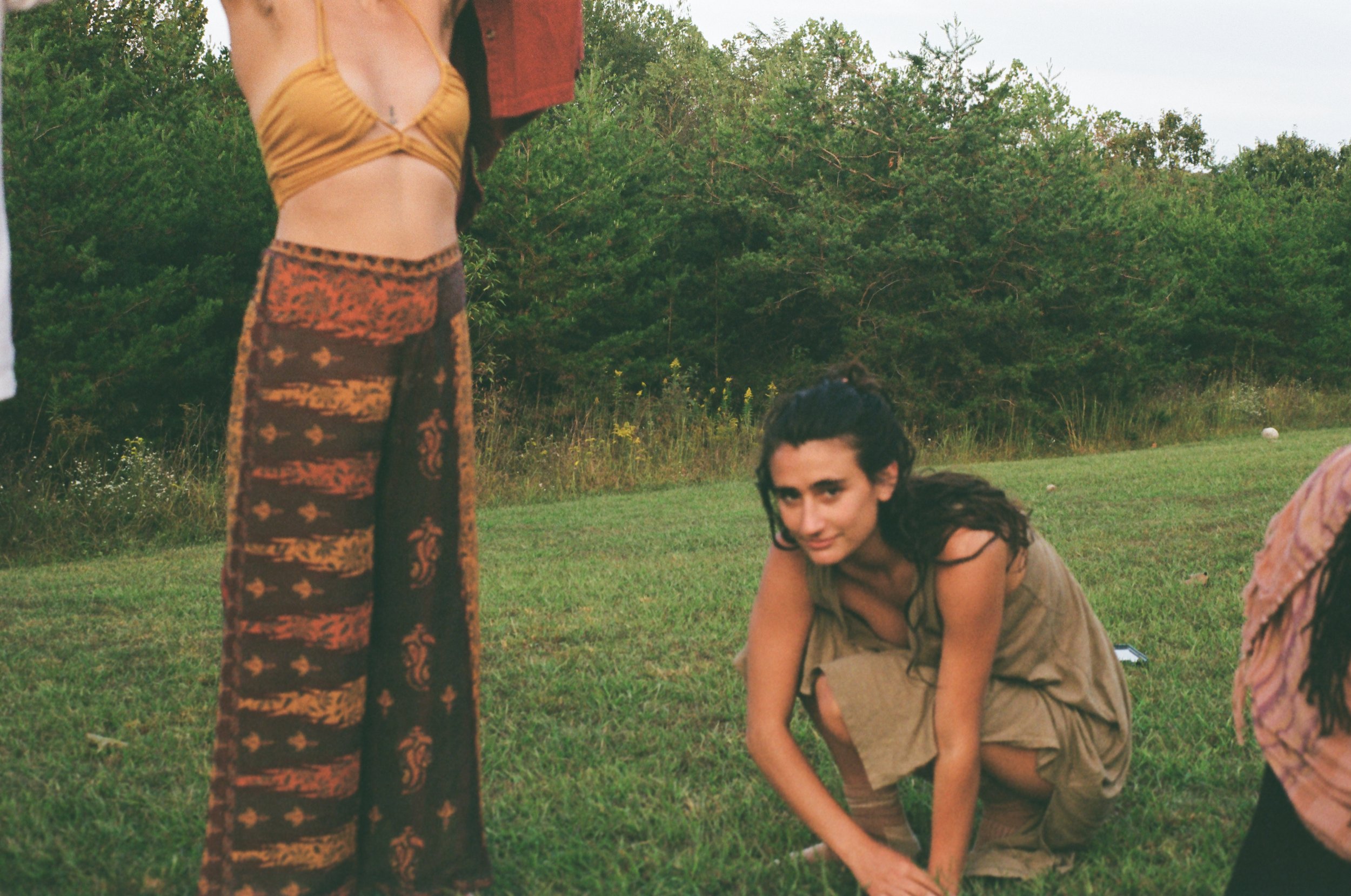 A young woman with long dark curly hair wearing a beige dress crouches on a grassy field surrounded by greenery. Partially visible on the left is another person wearing colorful, patterned wide-leg pants and a mustard yellow top, with the person's face not shown. The background features trees and wildflowers, and the overall setting appears to be outdoors in a natural environment.