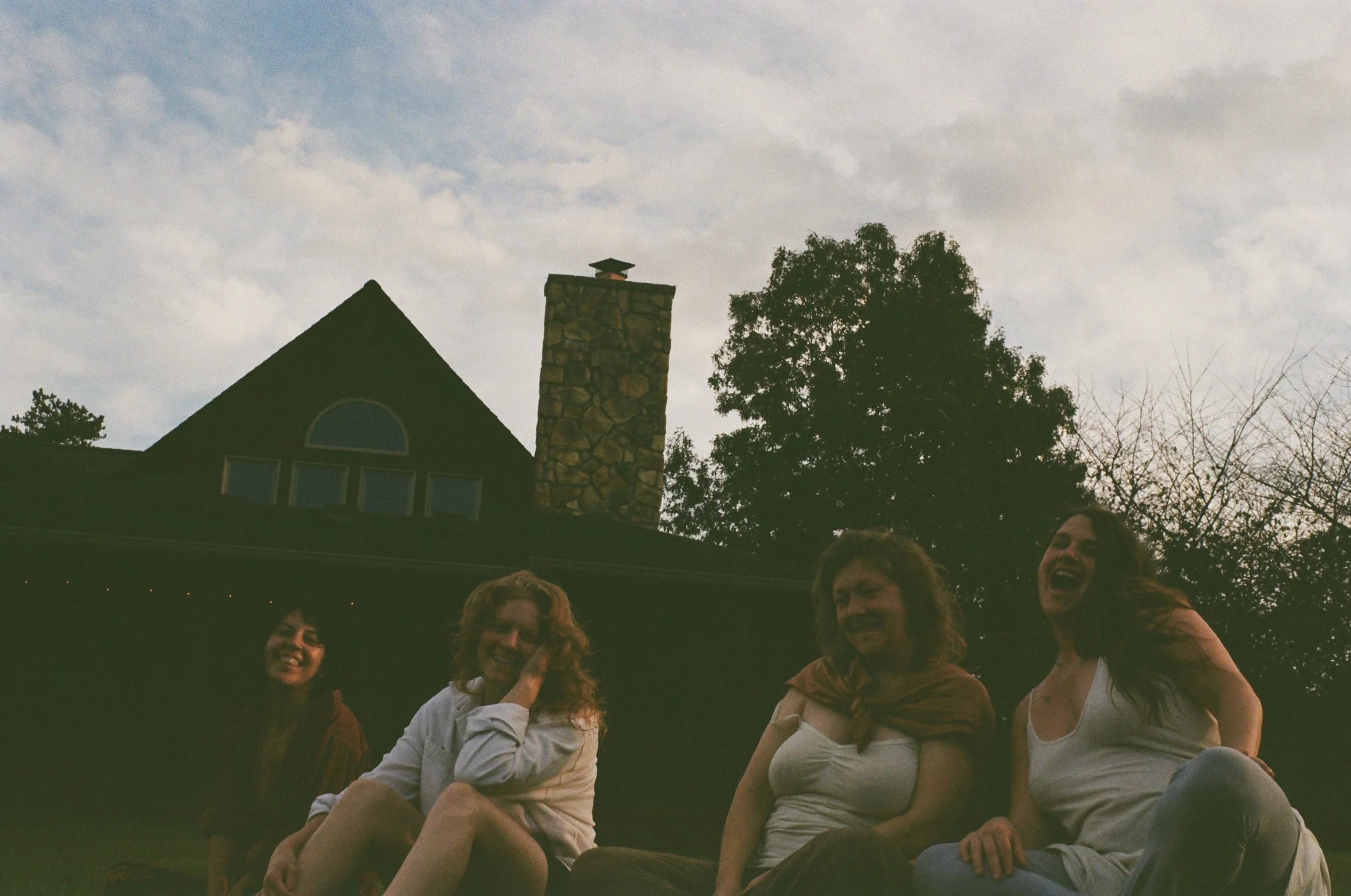 Four women sitting on grass outdoors, smiling and laughing, with trees, a house, and a cloudy sky in the background.