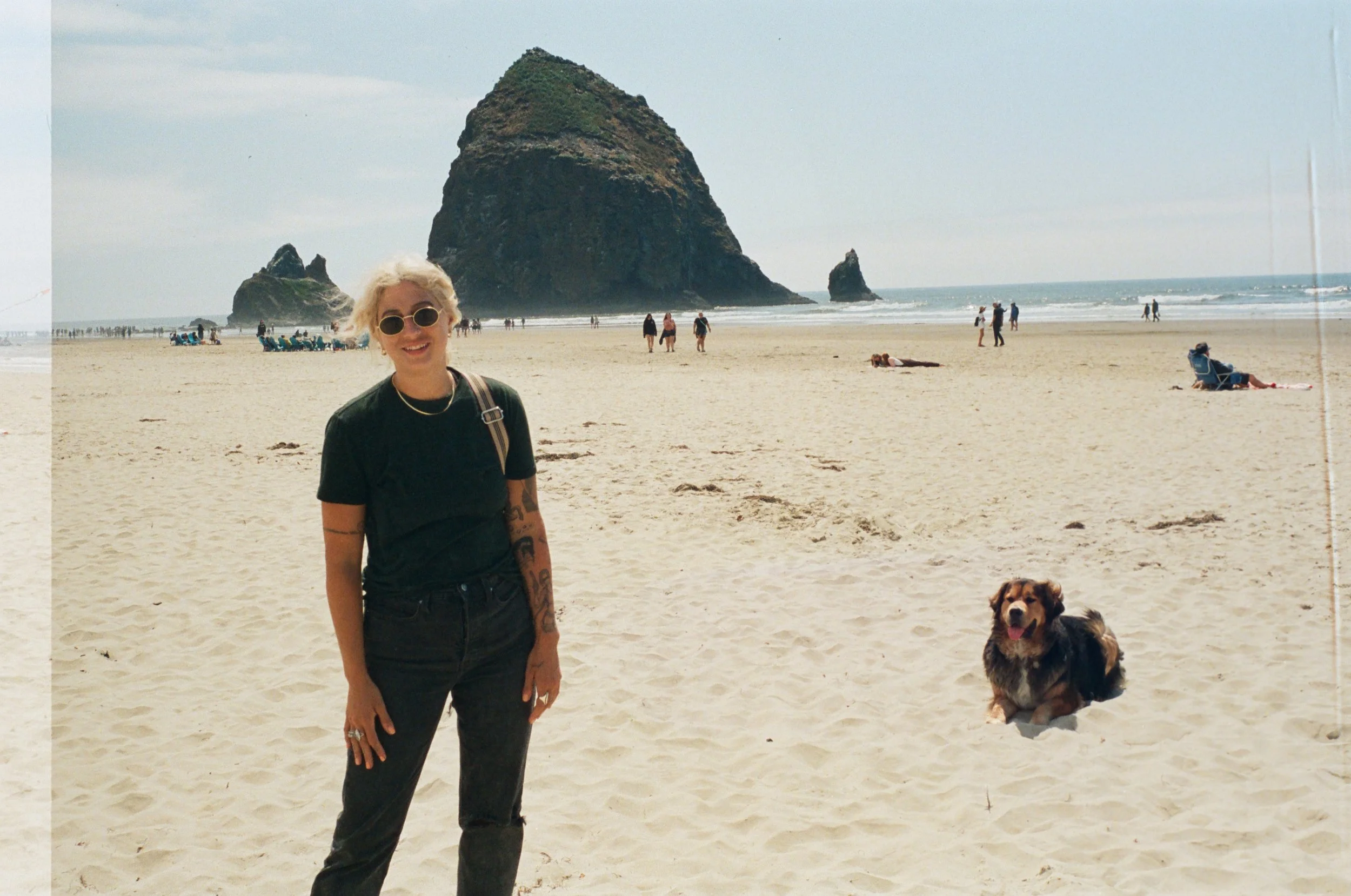 A woman with blonde hair, sunglasses, and tattoos standing on a sandy beach next to a dog with a background of large rock formations and people walking near the water.