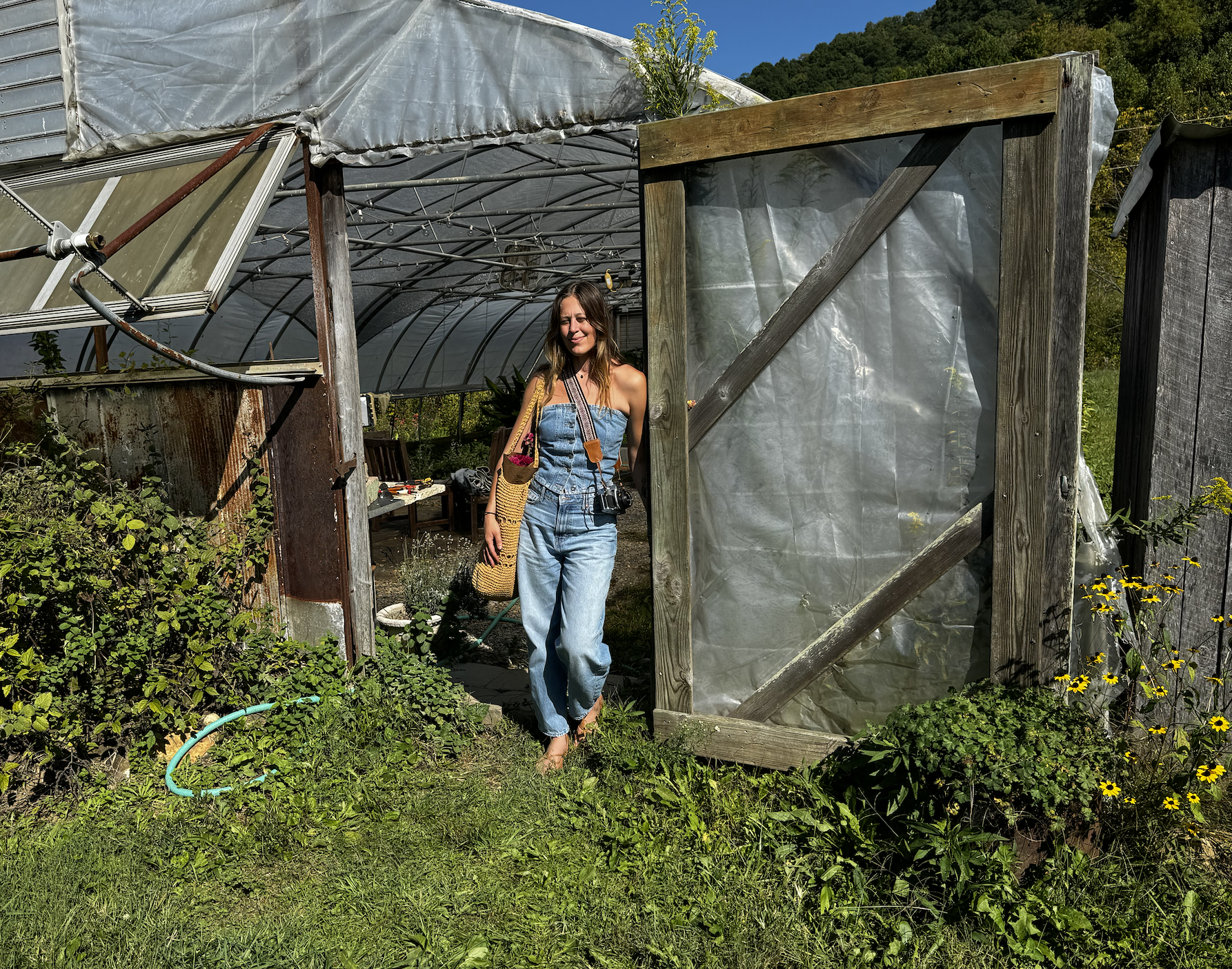 A woman in denim overalls and a strapless top standing in front of a greenhouse with various plants and garden furniture, surrounded by greenery and yellow flowers.
