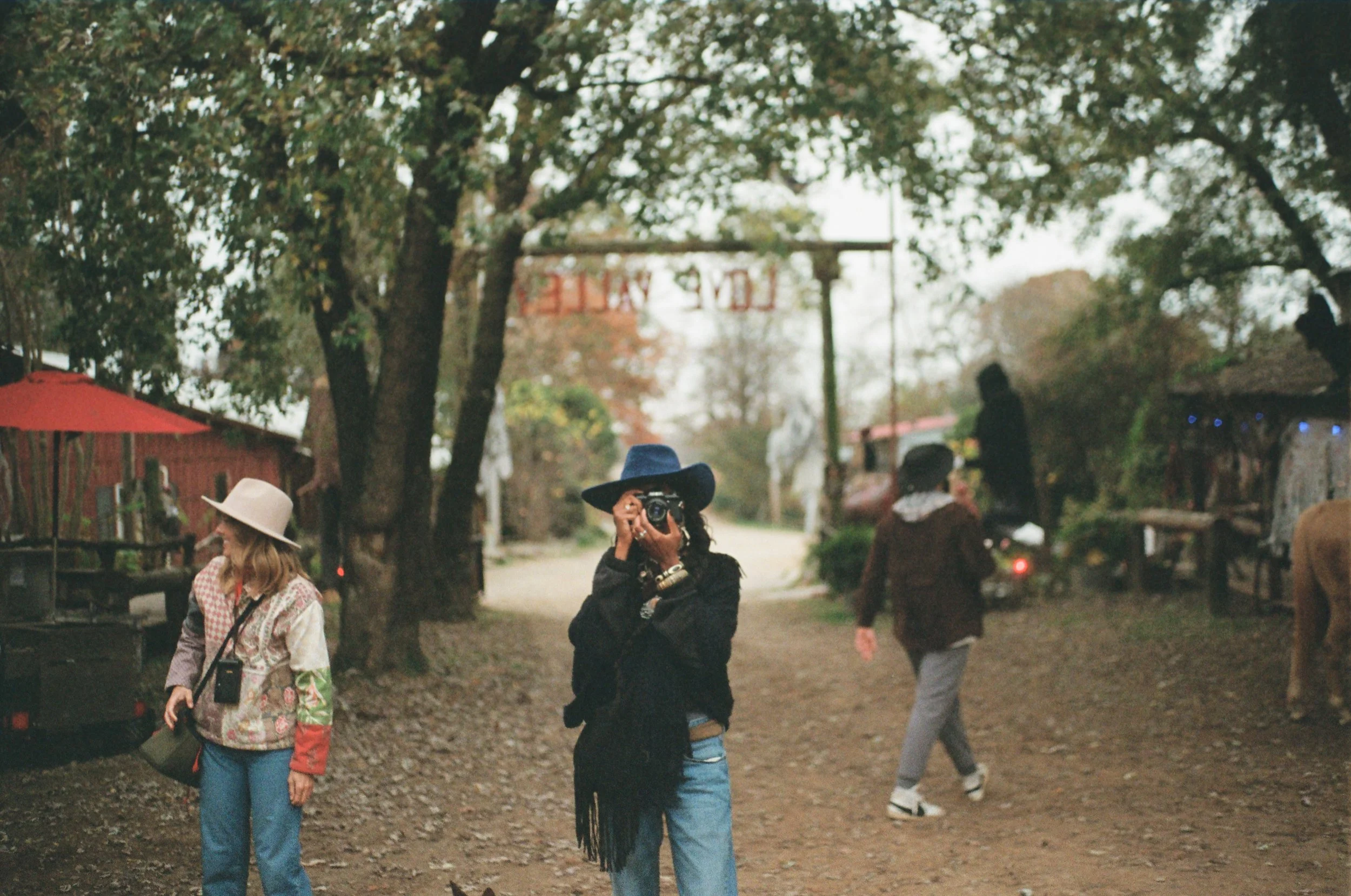 People outdoors in a wooded area with trees and a dirt path. One woman is taking a photo with a camera, wearing a wide-brimmed hat and blue shirt. Other individuals are walking or standing nearby.