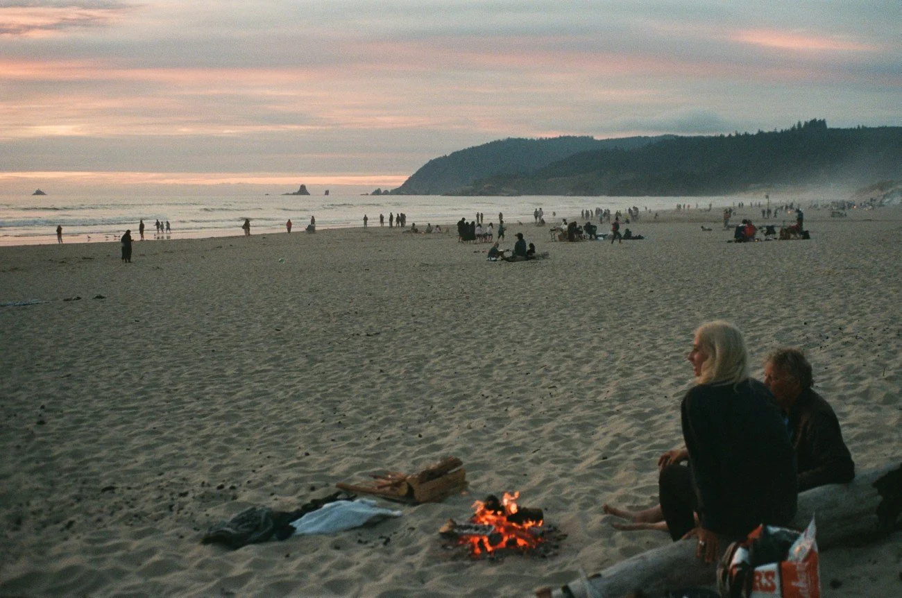 People gathered on a sandy beach during sunset, some sitting around a small campfire, with the ocean and cliffs in the background.