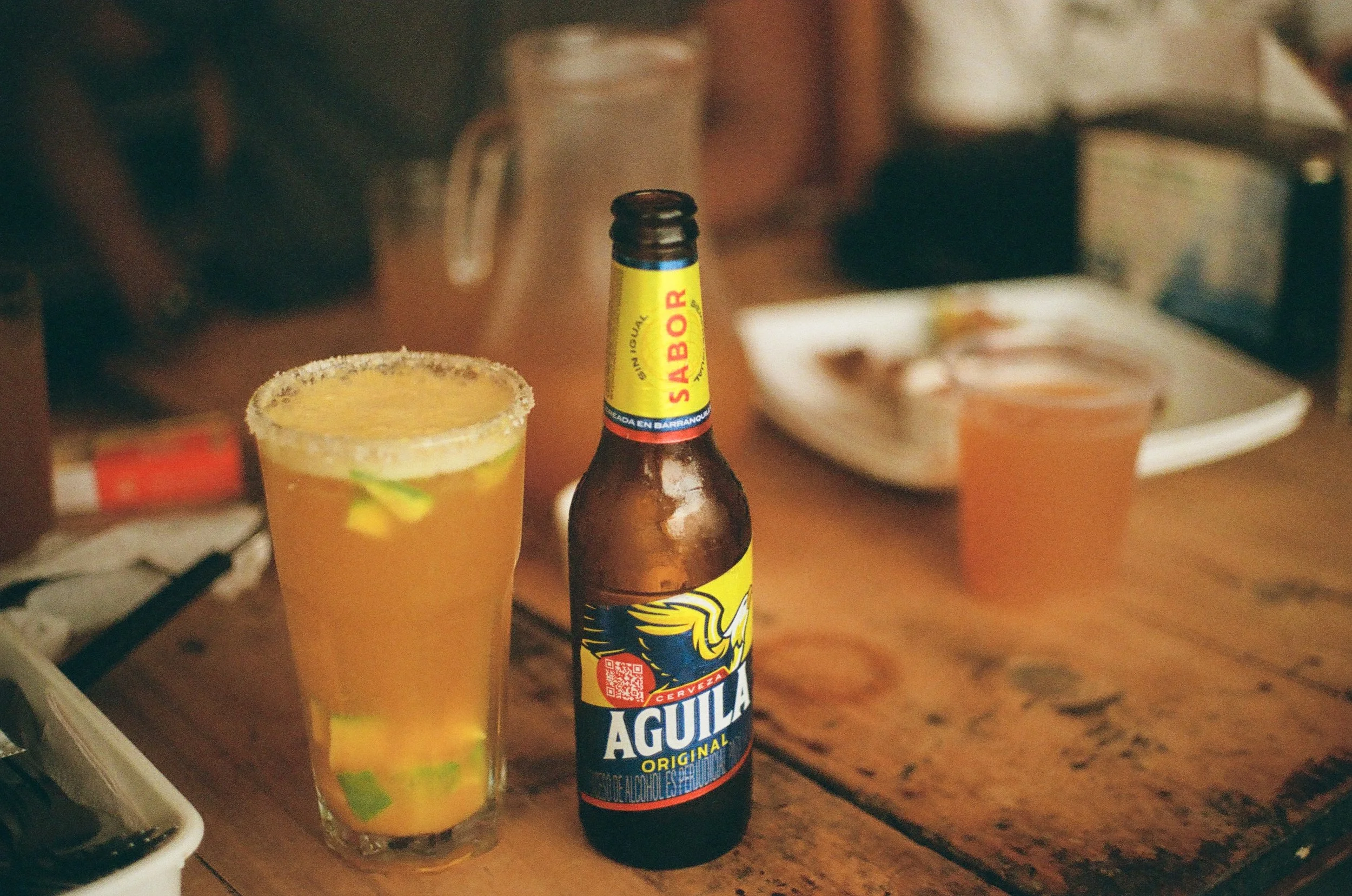 A glass of beer with a salted rim and lime slices and a beer bottle on a wooden table. In the background, a plate and another drink are visible.