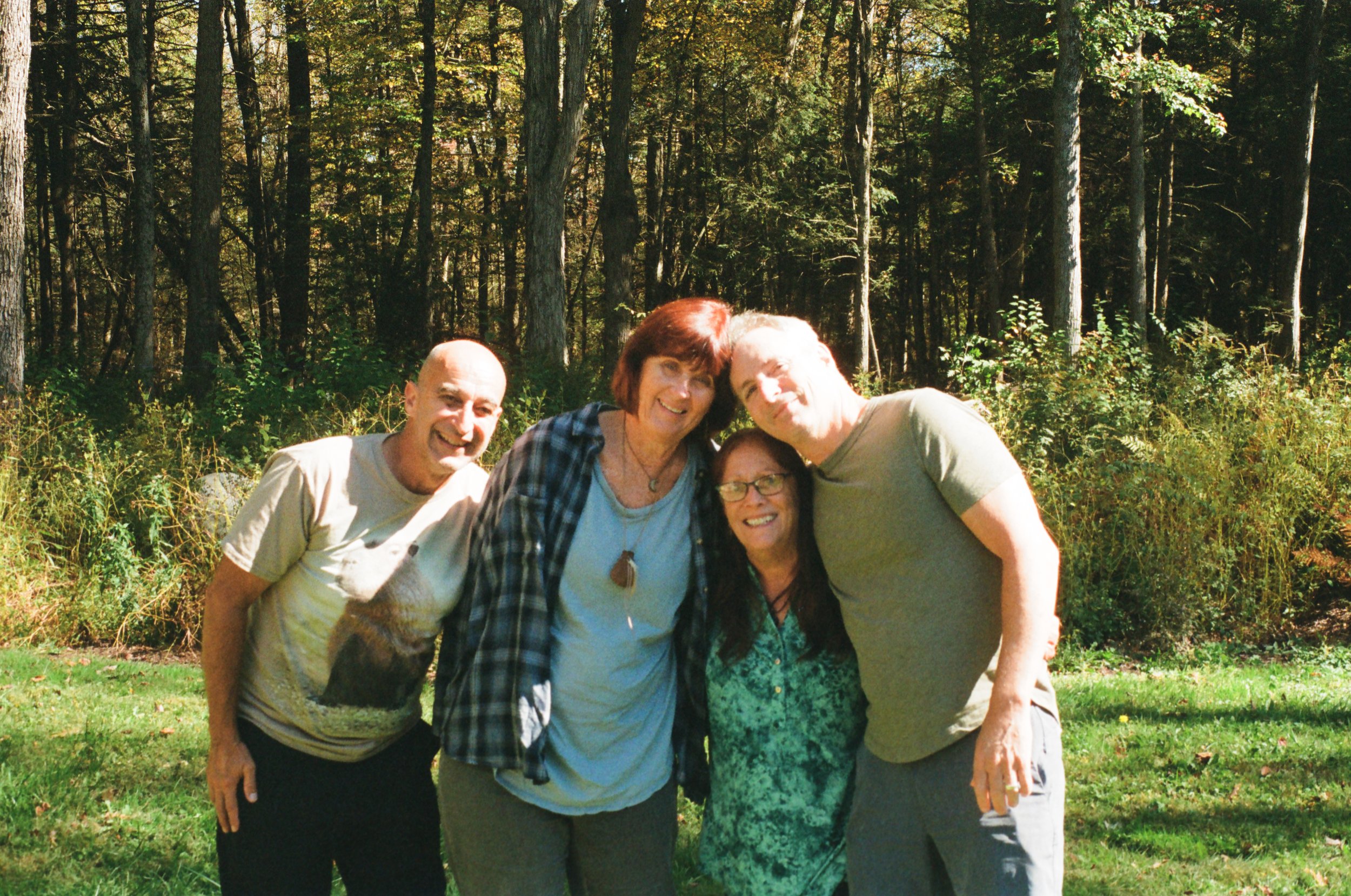 Four people outdoors in a forest, smiling. Two women and two men, standing close together with their arms around each other.