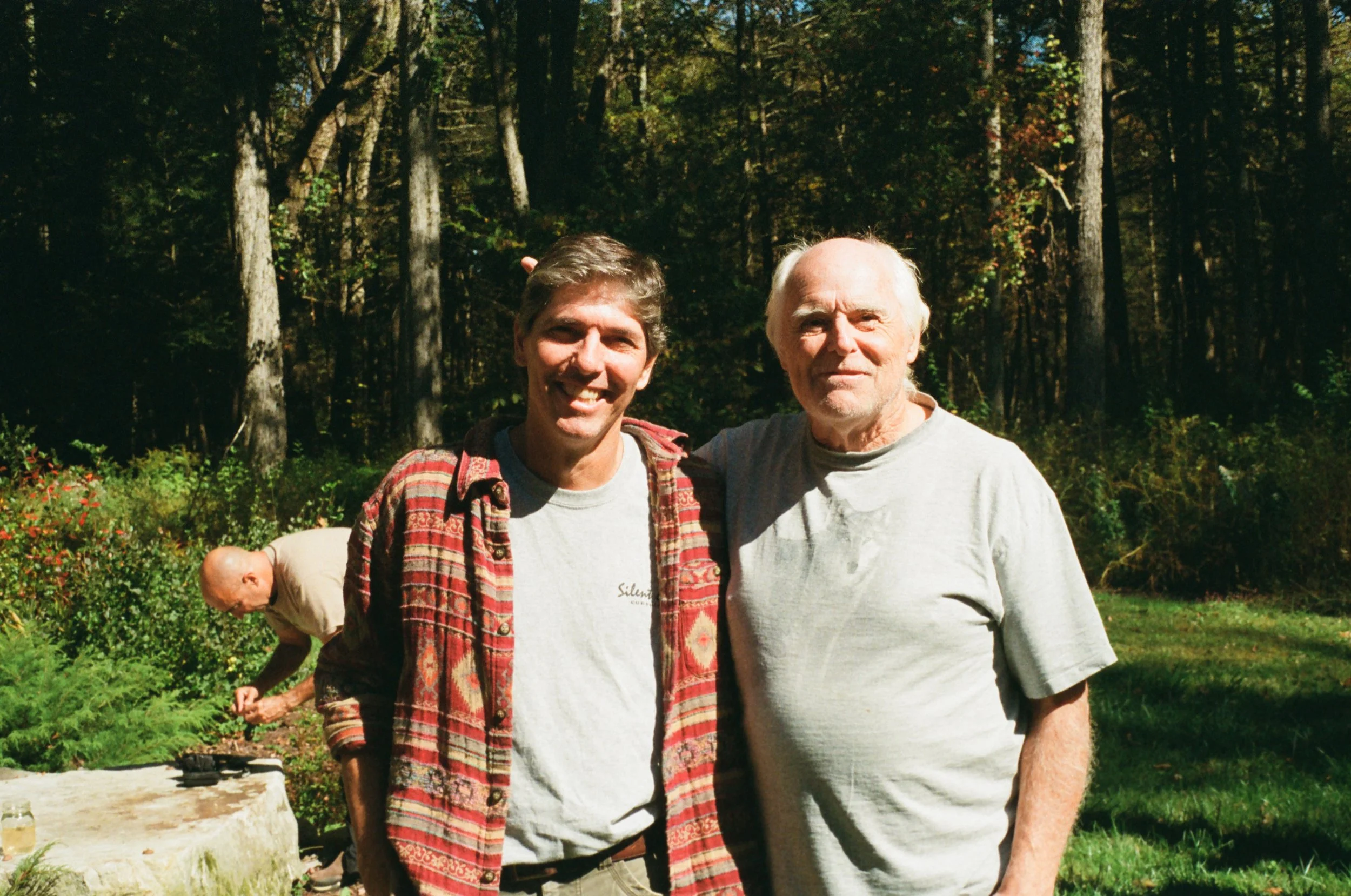 Two men standing outdoors in a forested area for a photo, with a person in the background gardening.