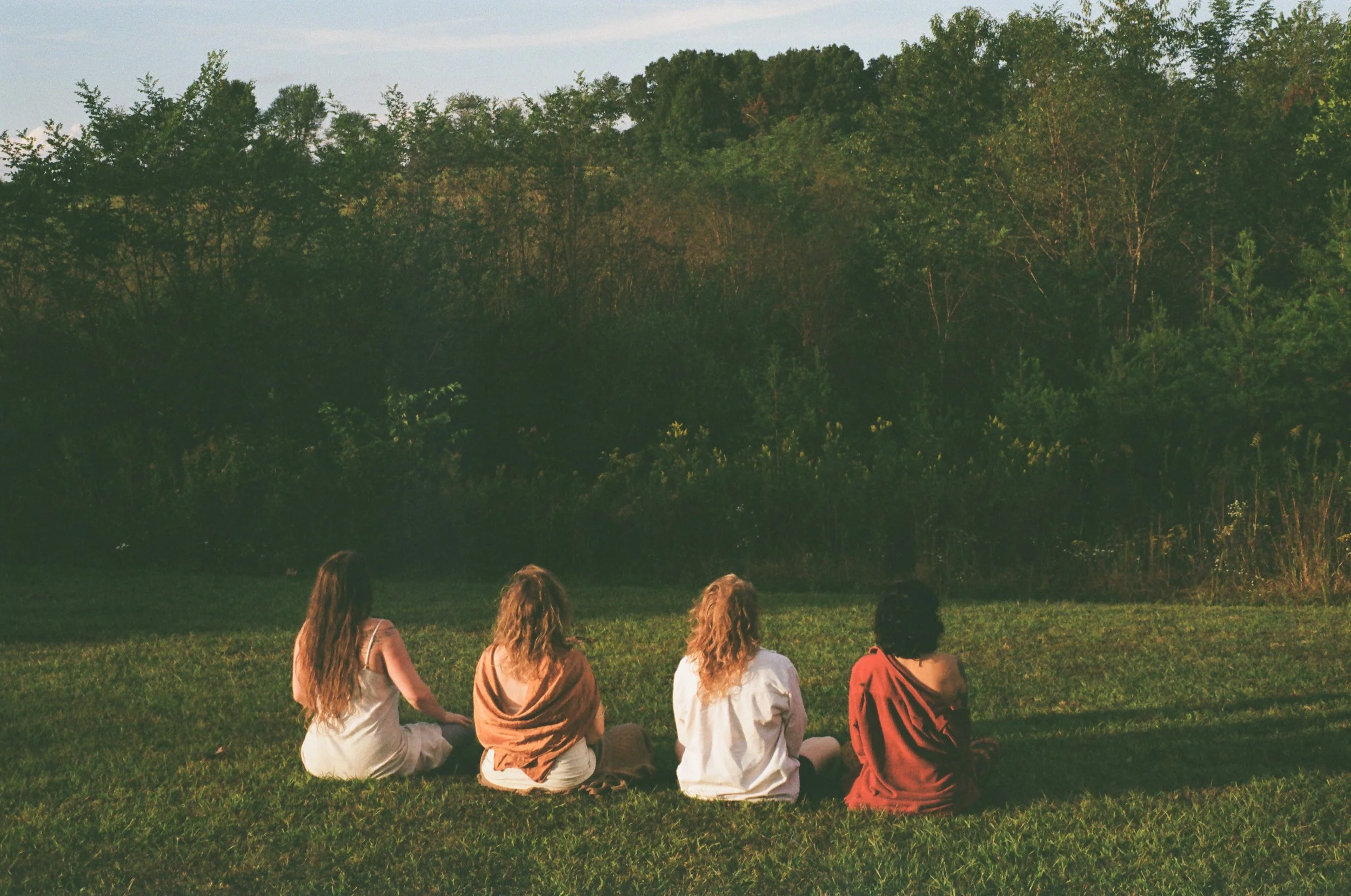 Four women sitting on the grass facing a wooded area during sunset.