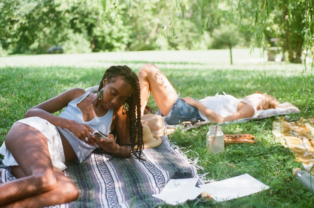 Two women relaxing on a grassy area with trees, one lying on a blanket using a phone and the other lying on a towel, surrounded by personal items like a jar, a hat, and portraits, in a park on a sunny day.