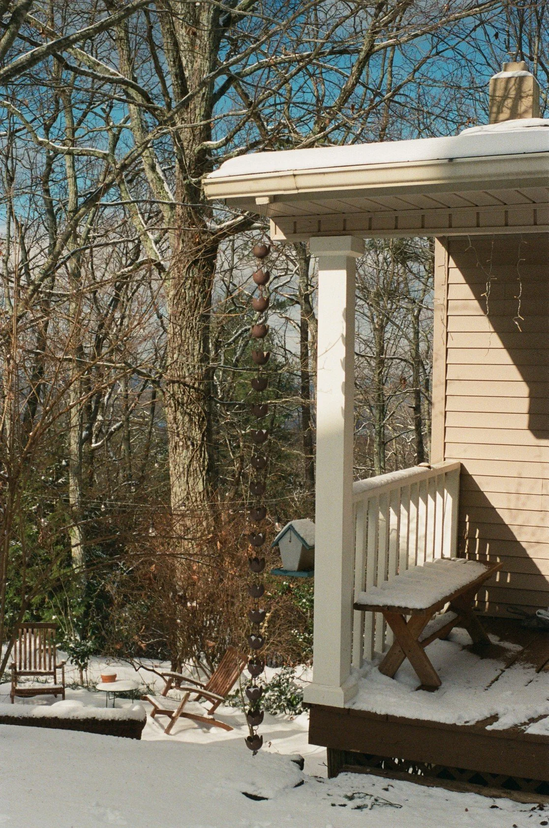 A backyard scene during winter with snow-covered ground, trees without leaves, a small porch with a bench, and a decorative hanging metal wind chime.