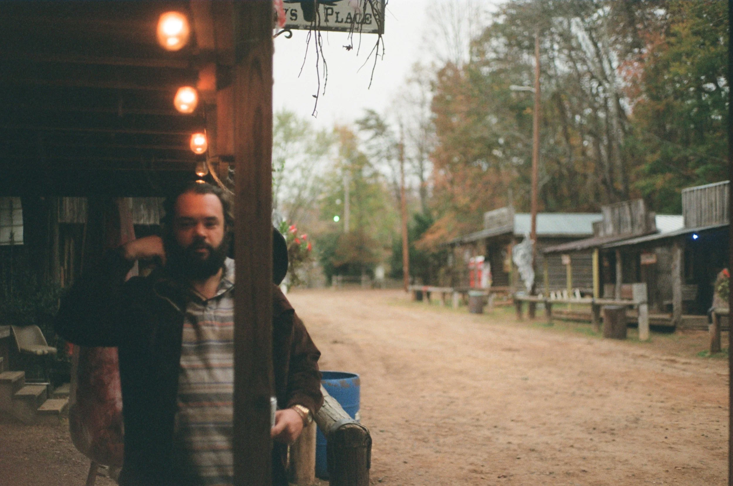 A man with a beard wearing a striped shirt and dark jacket stands under a wooden structure decorated with string lights, outdoors in a rustic setting with a dirt pathway and wooden buildings.