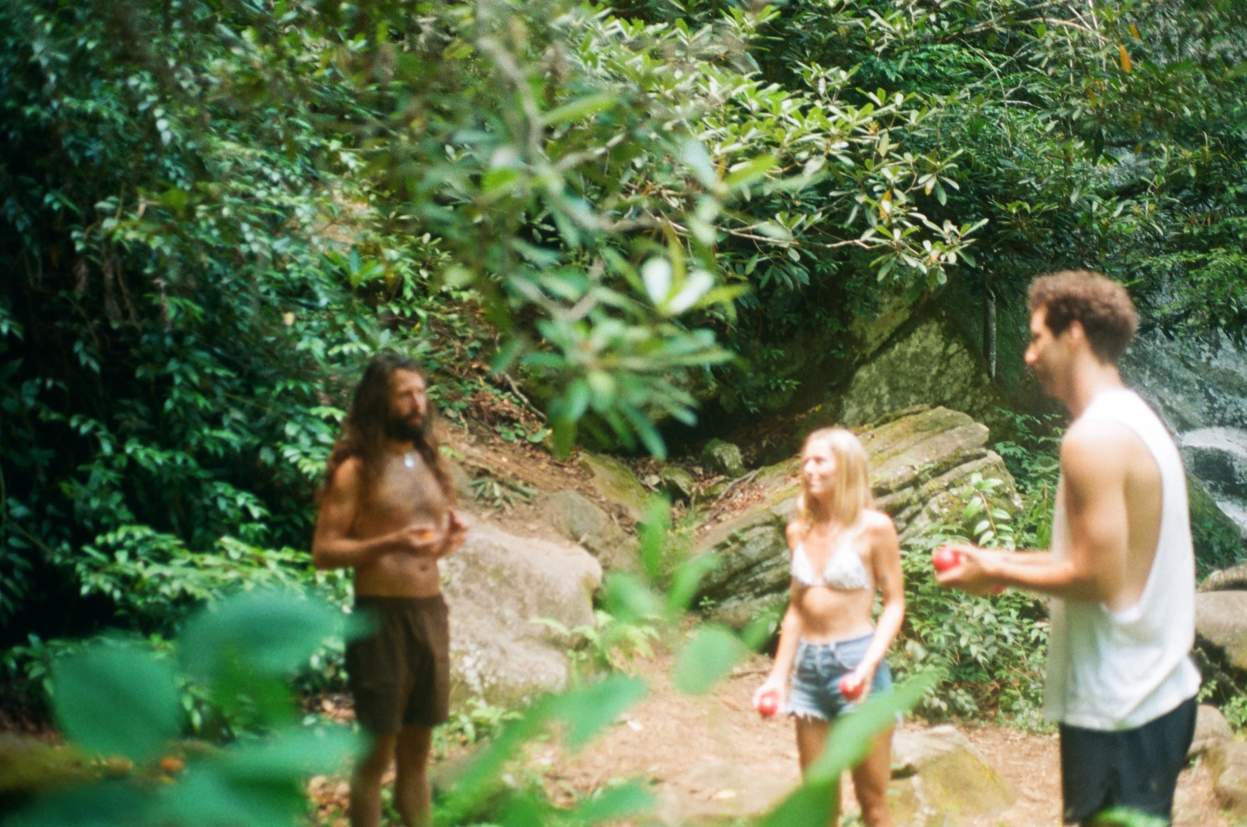 Three people, two men and one woman, in a forest clearing surrounded by dense green foliage and rocks. One man is shirtless with long hair and a beard, holding a drink. The woman has blonde hair, is wearing a white bikini top and denim shorts, and is holding red balls. The other man is wearing a sleeveless white shirt and black shorts, holding a red ball.