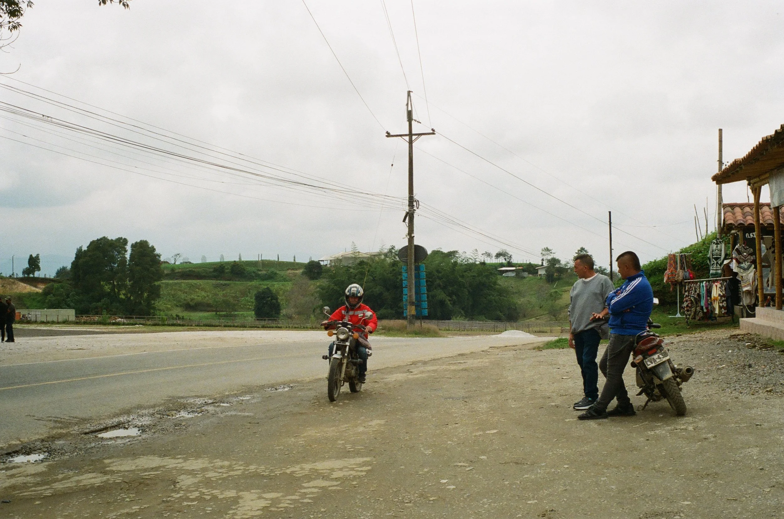 Two men standing on a gravel roadside next to a motorcycle, with a person riding a motorcycle approaching them on a paved road. There are small shops or stalls on the right, and electrical poles and wires overhead. Green hilly landscape in the backgr