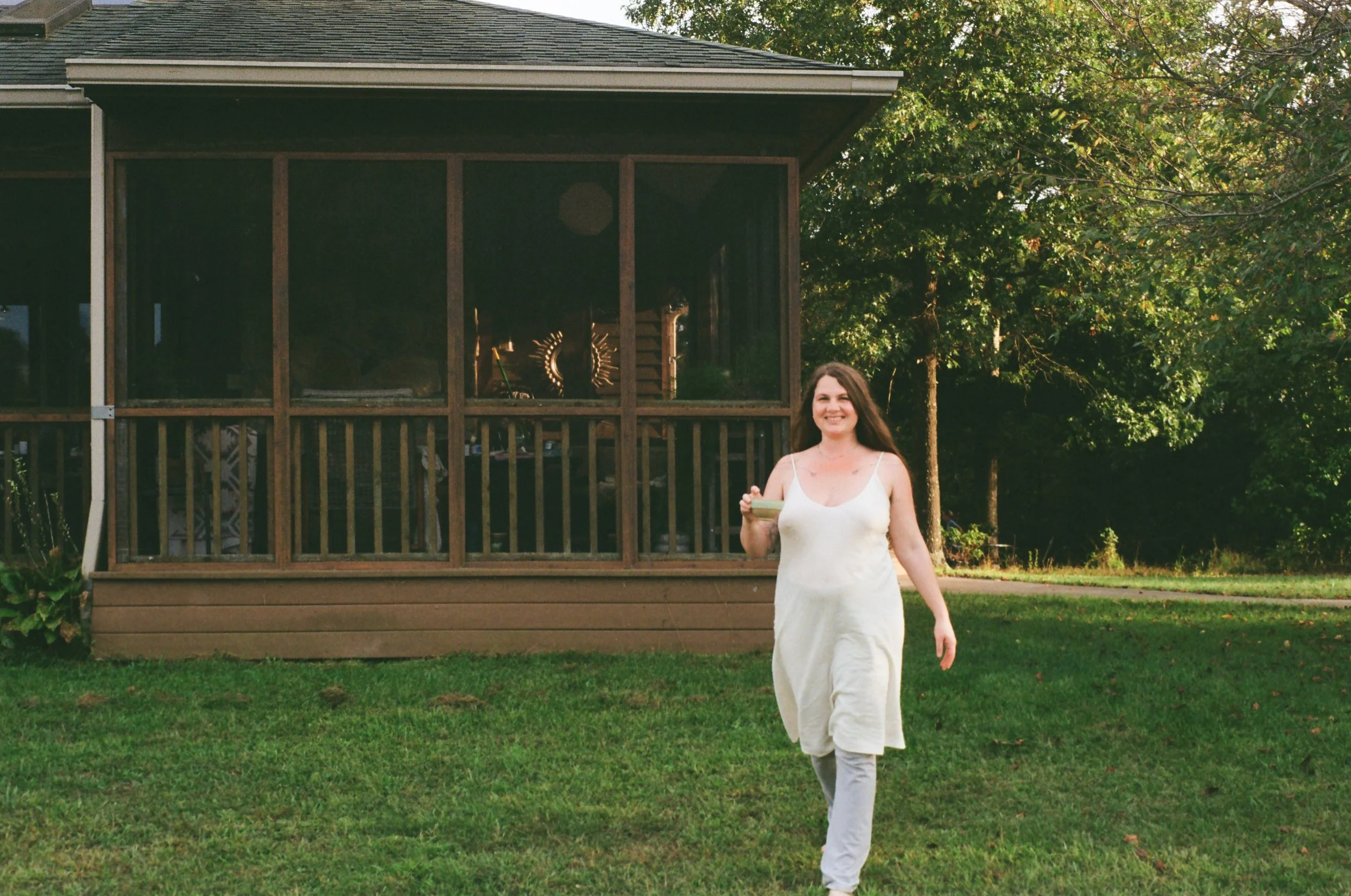 A woman in a white dress and leggings walking on a green lawn holding a small container with a spoon, smiling. In the background, there is a house with a screened porch and trees.