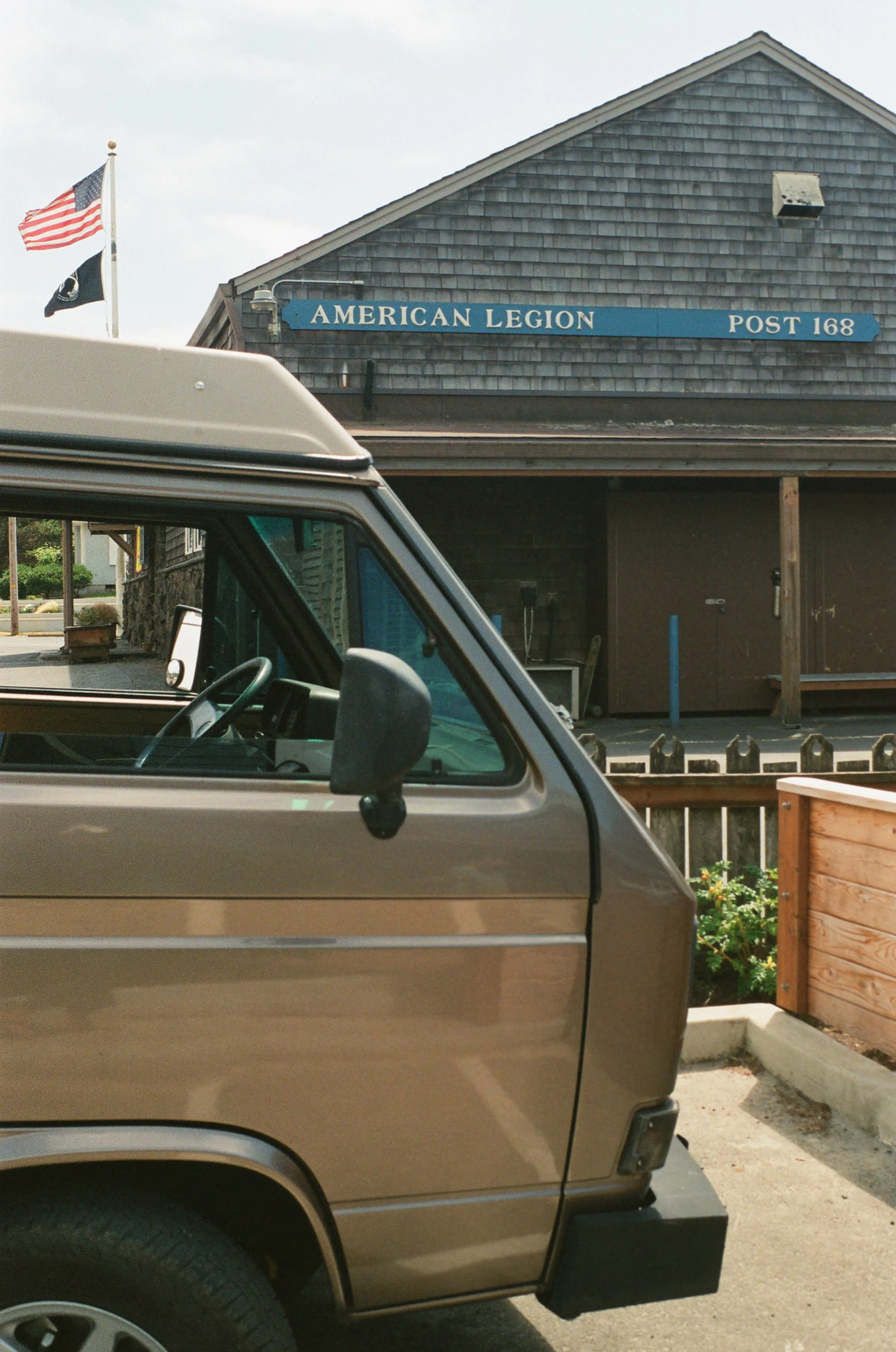 Partial view of a vintage beige pickup truck parked outside a building with a sign that reads 'American Legion POST 168'. American flags are flying nearby.
