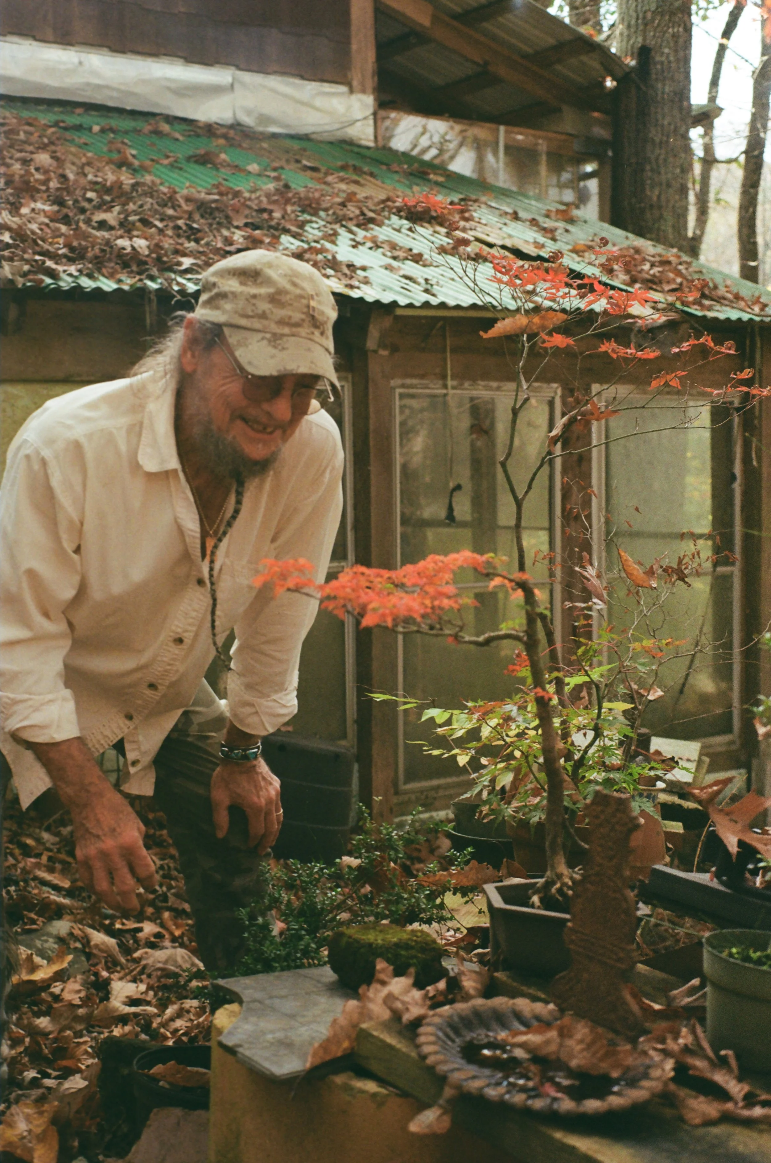 An elderly man with glasses, a beige cap, and a light shirt, smiling and leaning over in a garden surrounded by autumn leaves and potted plants, near a rustic building with a sheet metal roof.