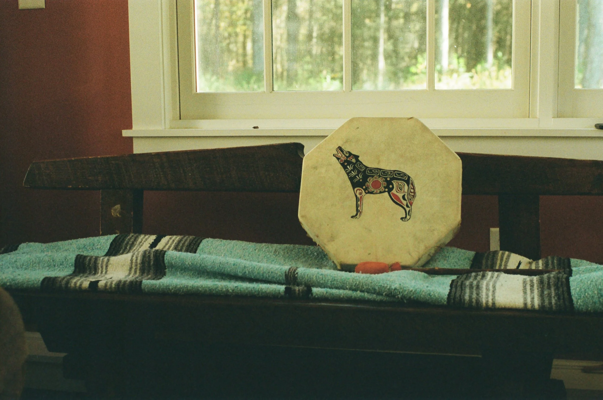 A wooden bench with a turquoise striped textile covering, positioned in front of a window with a forest view outside. On the bench, there is an octagonal painted stone featuring a stylized black cow with red and white details.