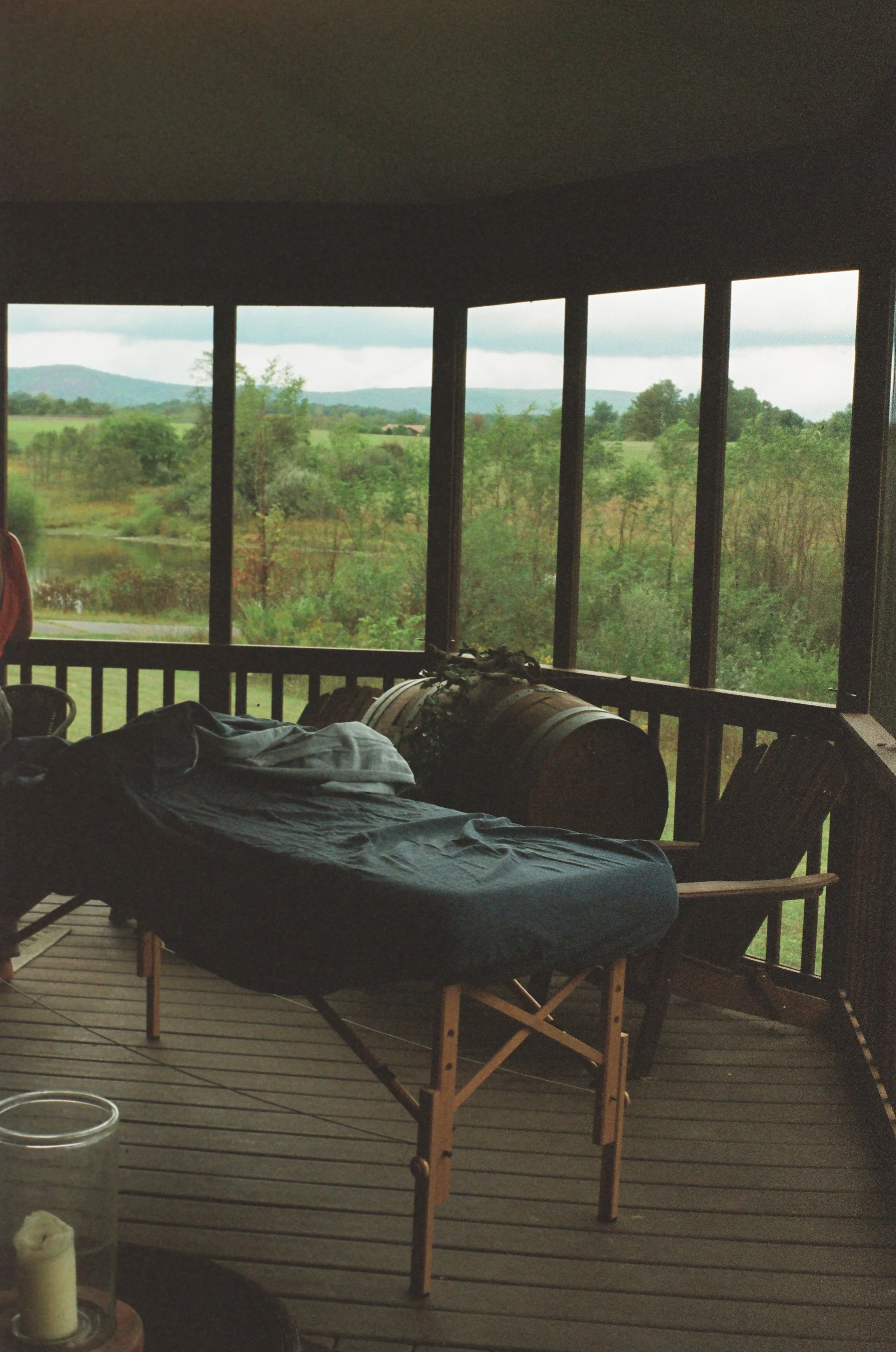 A screened porch with a massage table, a large barrel, and outdoor scenery with green trees, grass, and distant hills under a cloudy sky.