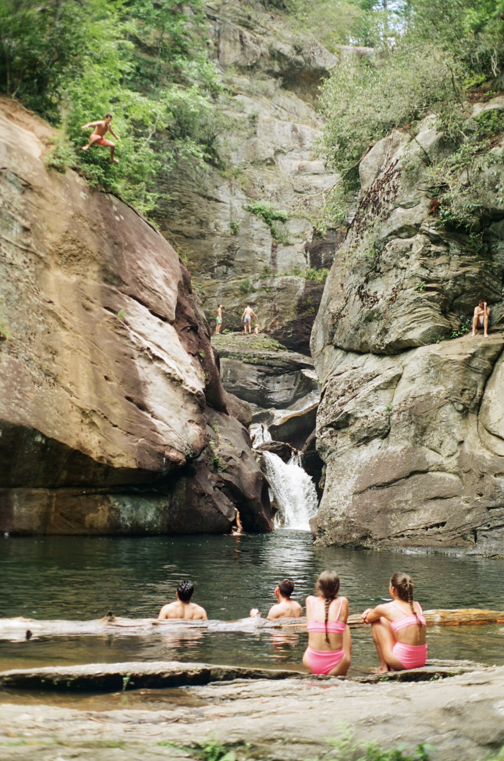 People swimming and relaxing near a waterfall in a rocky canyon surrounded by green trees.