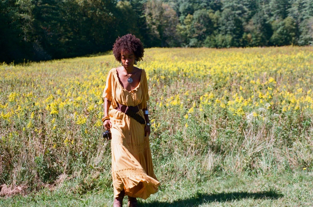 A woman in a yellow dress walking through a field of yellow flowers with a forest in the background.