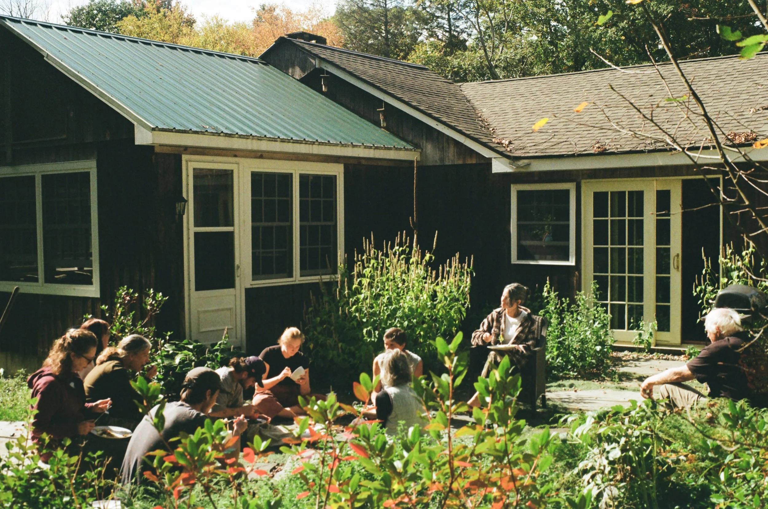 Group of people sitting on the grass in a garden breakfast or brunch, with a woman standing and speaking, near dark-colored houses with large windows, surrounded by greenery.