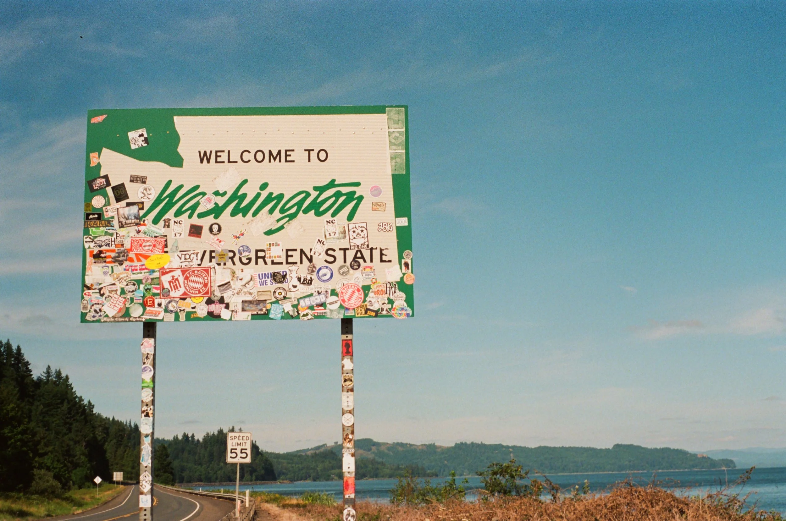 A highway sign in Washington state covered with numerous stickers, with a scenic landscape of water, trees, and hills in the background under a blue sky.
