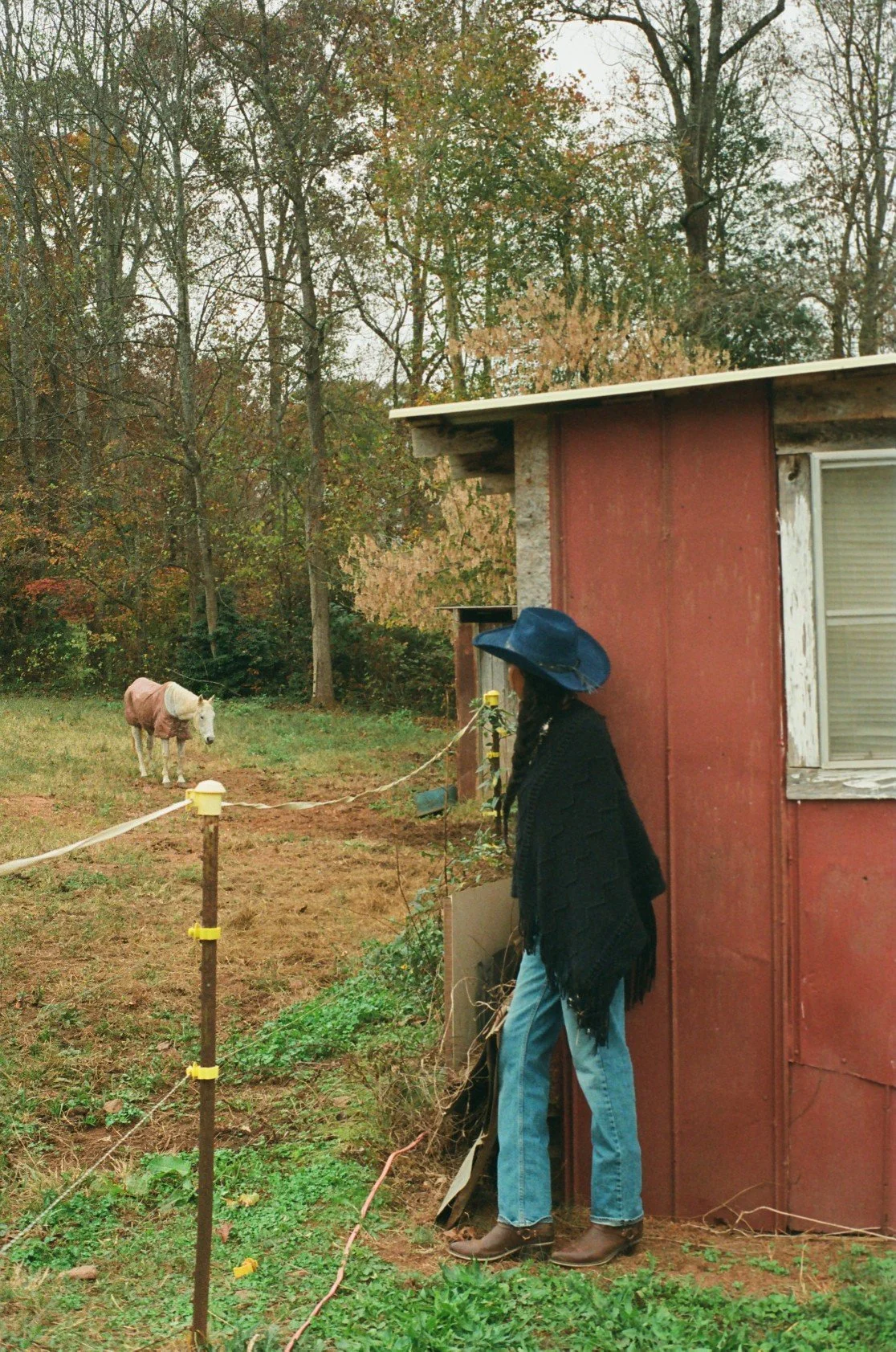 A woman wearing a black poncho, blue jeans, cowboy boots, and a wide-brimmed blue hat, standing outside a red wooden building. She is looking at a donkey in a fenced yard. The donkey is wearing a pink blanket, and there are trees with autumn foliage 