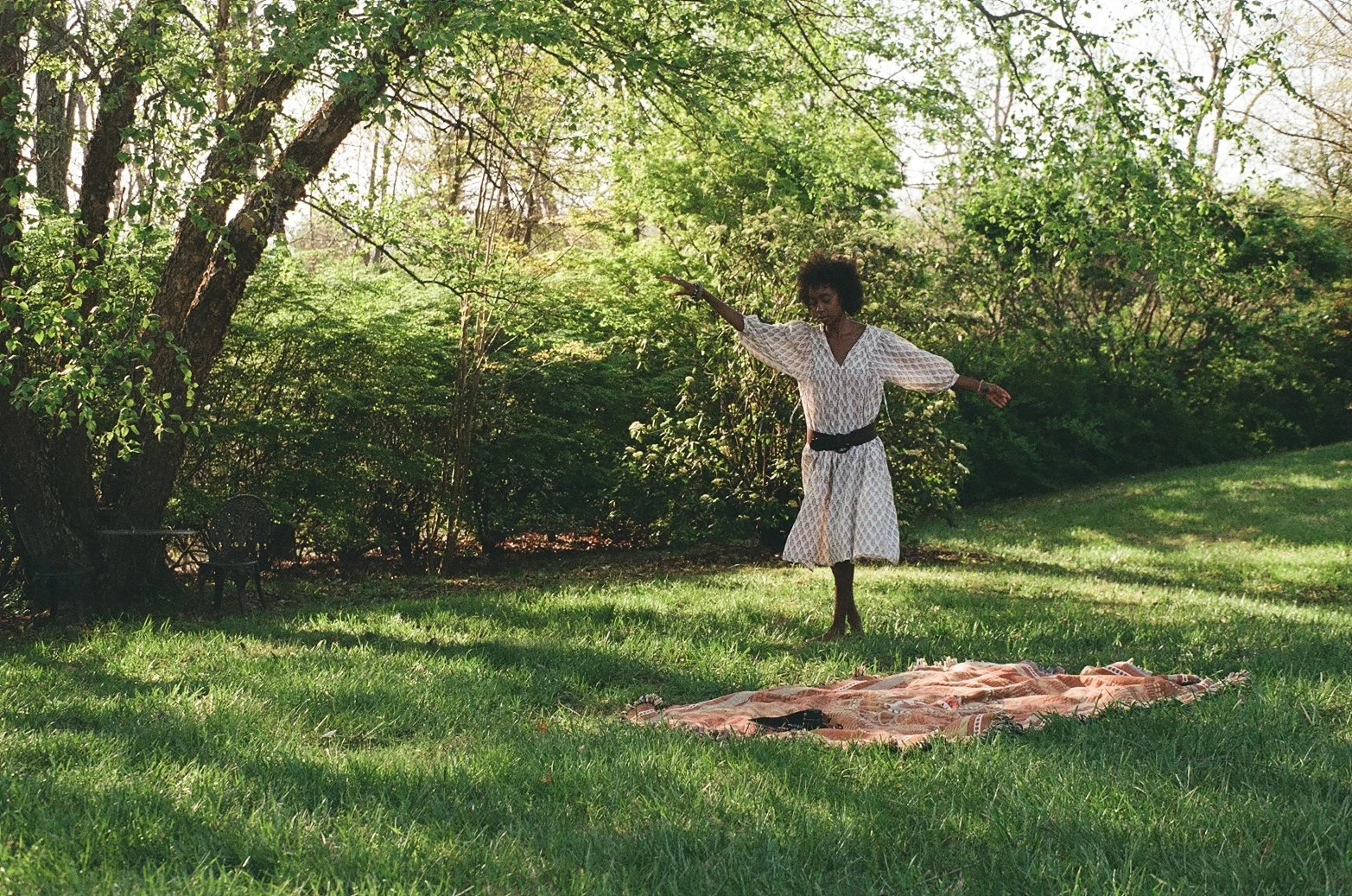 A woman in a white dress with a black belt, standing on one leg with arms outstretched, on a grassy lawn with trees and bushes in the background, and a blanket on the ground.