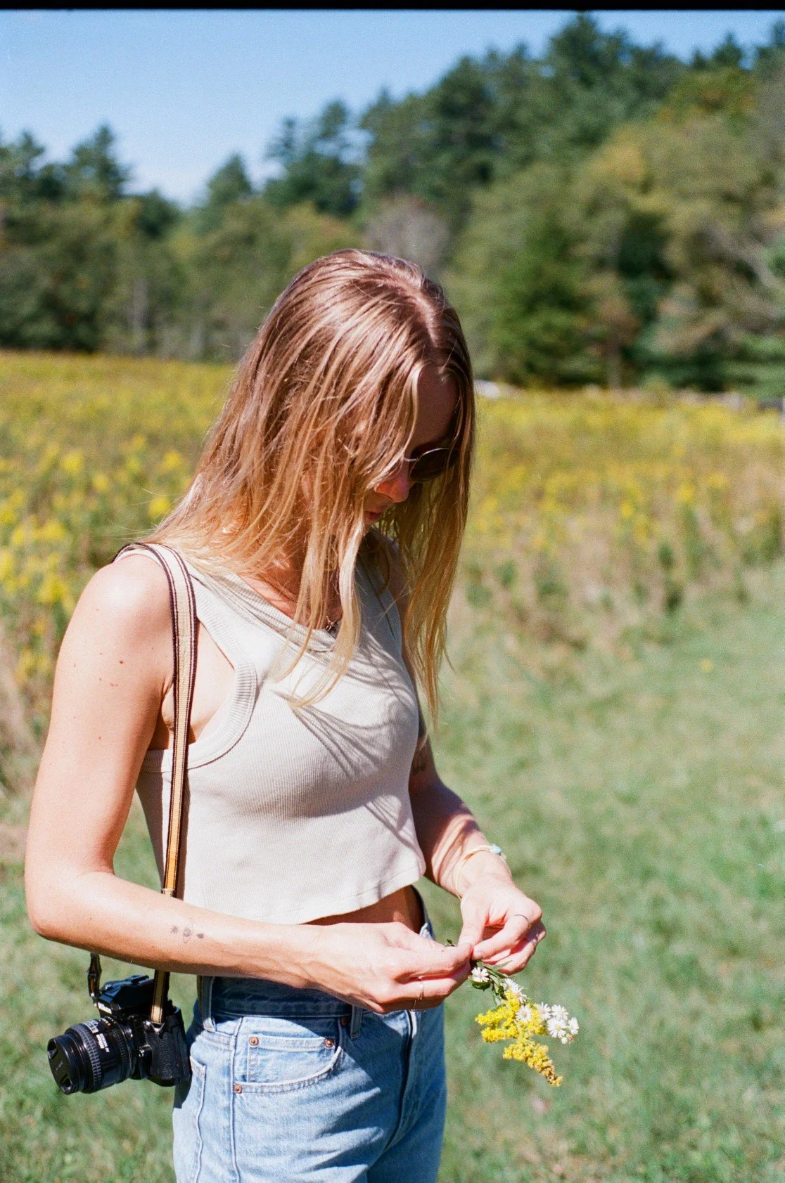 A young woman with long blonde hair, wearing sunglasses, a sleeveless beige top, and light blue jeans, stands outdoors in a grassy field with a camera hanging around her neck. She is holding a small bunch of flowers and looking down at them.