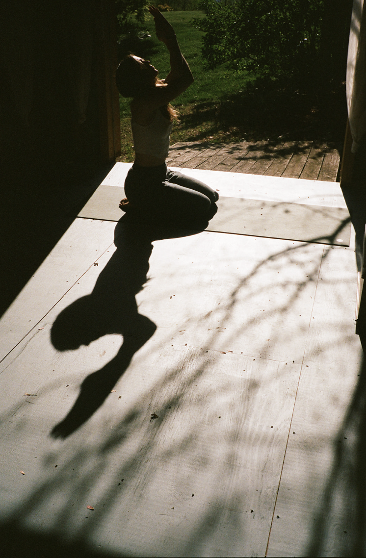 Person kneeling on a porch, arms raised towards the sunlight, with their shadow cast on the wooden floor and greenery visible outside.