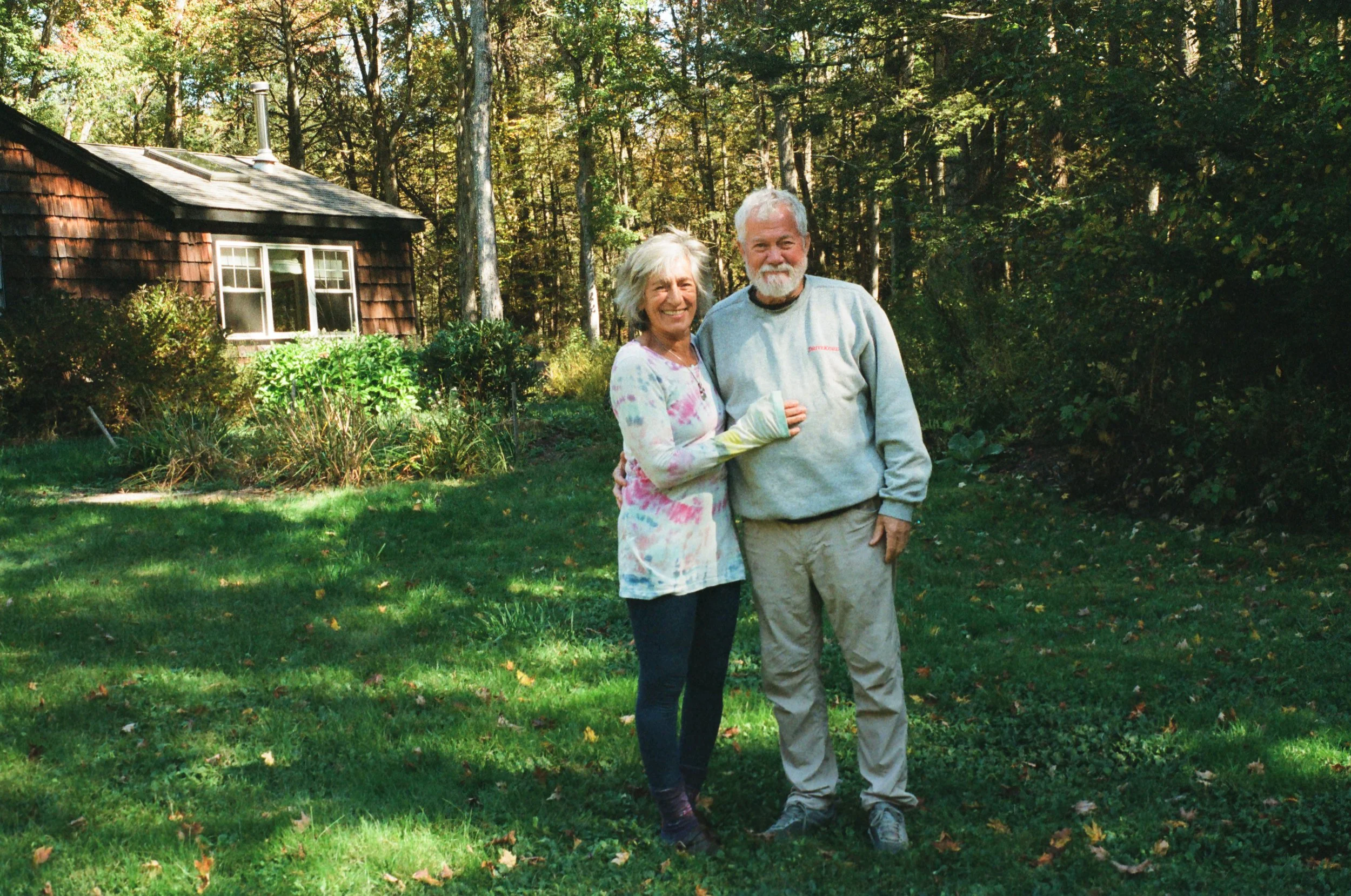 An elderly woman and man standing together on a grassy yard with trees in the background. The woman is smiling, wearing a multicolored long-sleeve top and dark pants. The man has a beard, is smiling, wearing a gray sweatshirt and light-colored pants.
