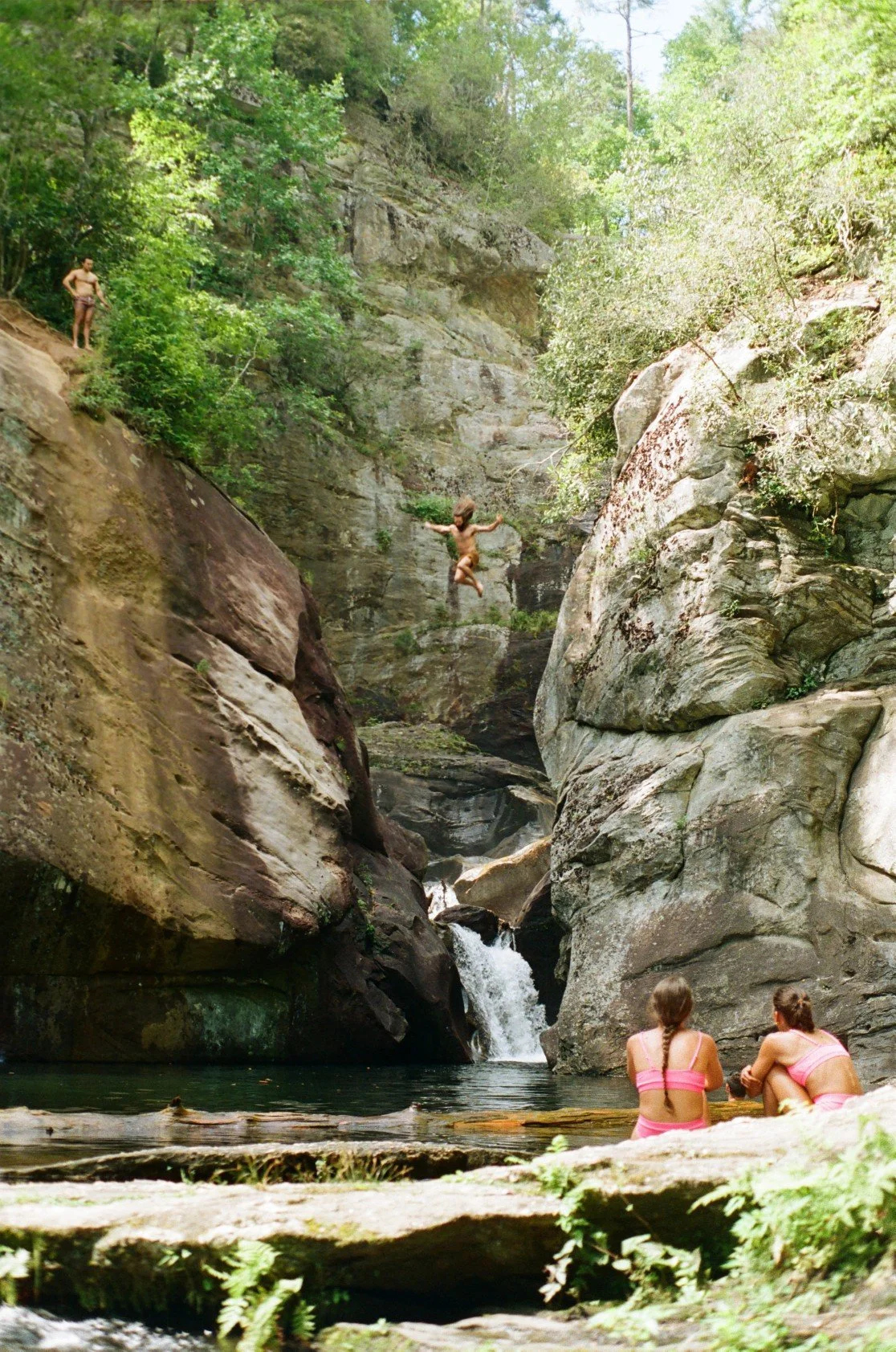 Two girls in pink swimsuits sitting by a stream watching a boy jump into the water from a rocky cliff in a lush, green canyon.