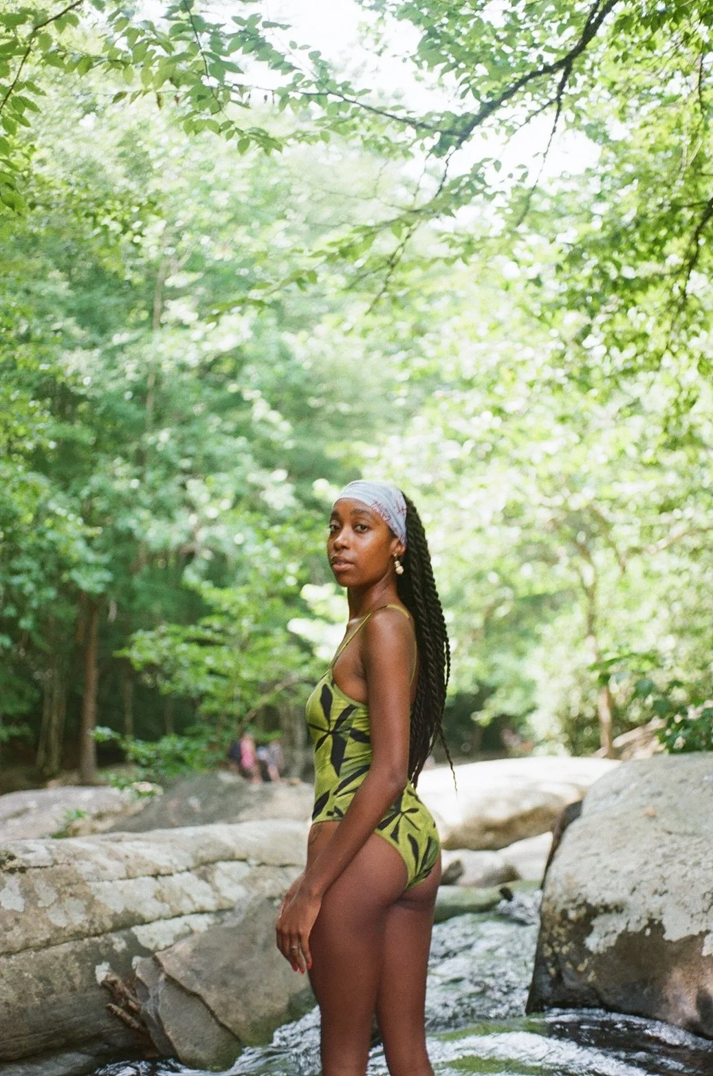 A woman in a green and black swimsuit with a light headscarf stands in a rocky stream surrounded by lush green trees.