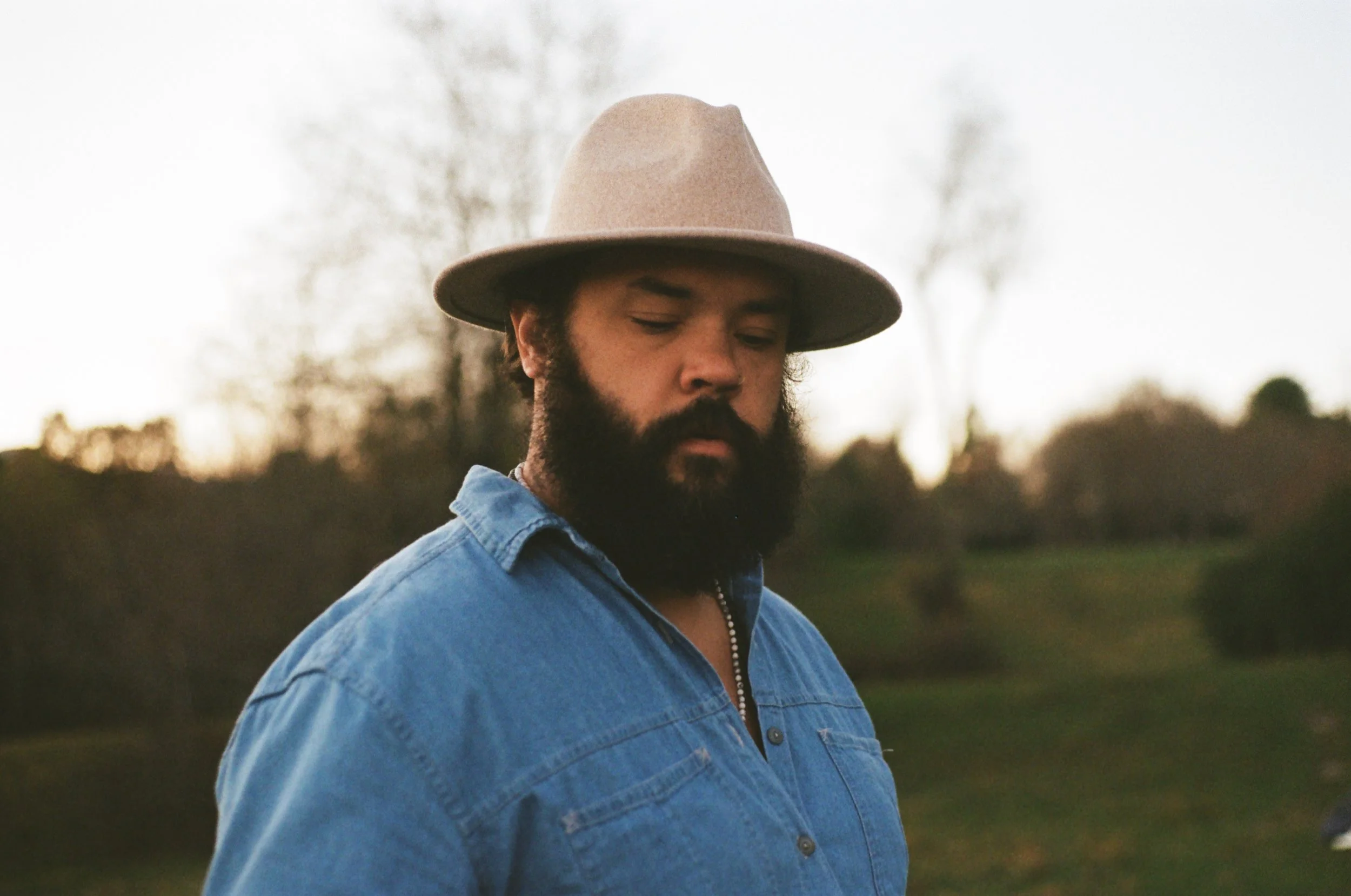 A man with a beard and mustache wearing a light brown wide-brimmed hat and a blue denim shirt, looking down outdoors during sunset in a park or natural setting with trees in the background.