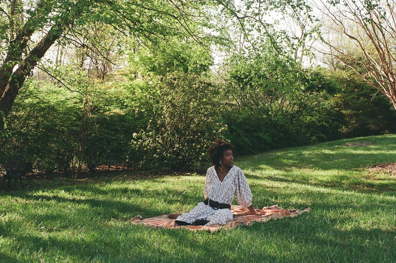 A woman sitting on a blanket on the grass in a park, surrounded by trees and bushes, with sunlight filtering through the leaves.
