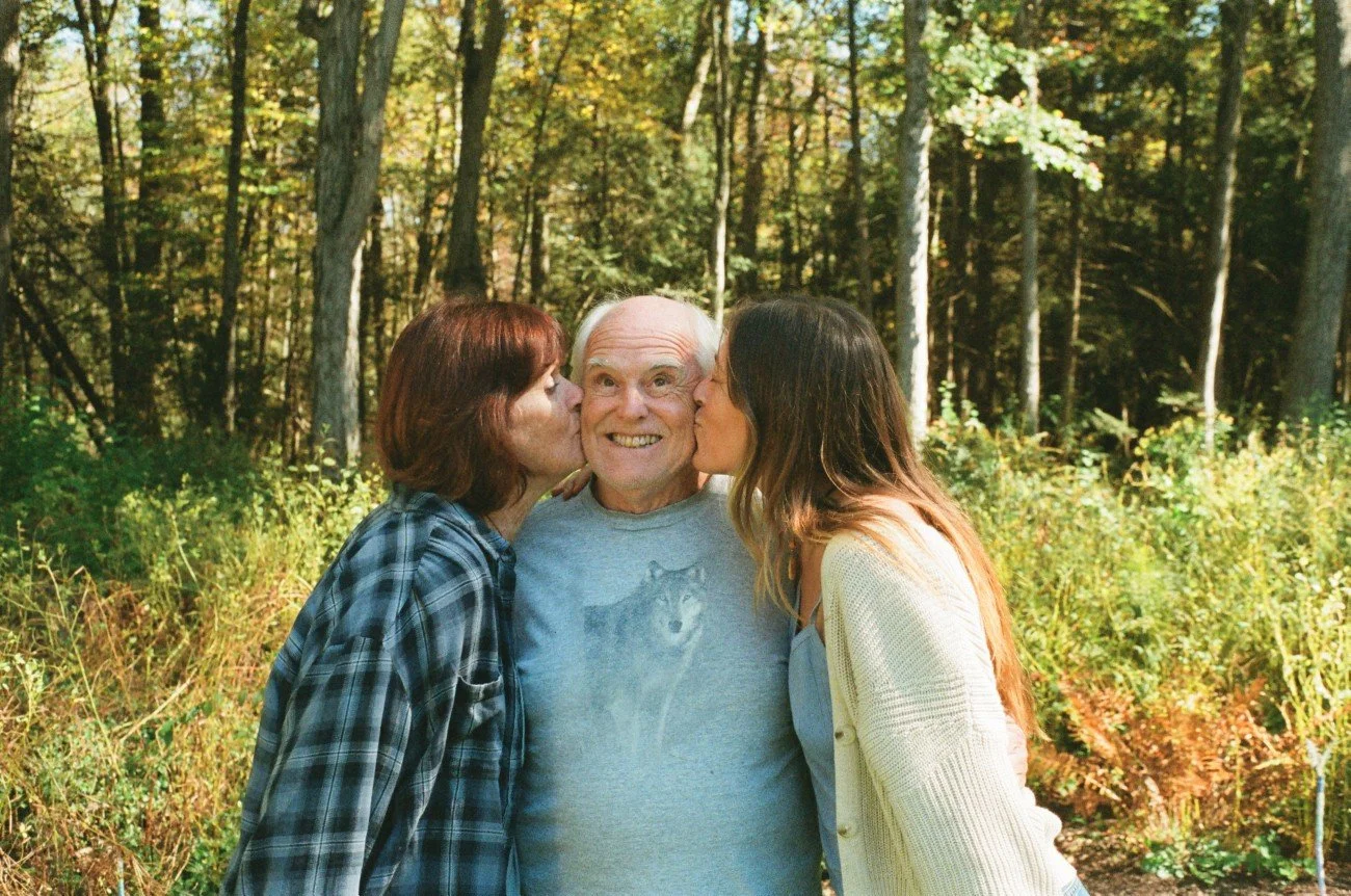 A man in a gray t-shirt with a wolf graphic is kissed on both cheeks by women on either side in a wooded outdoor setting.