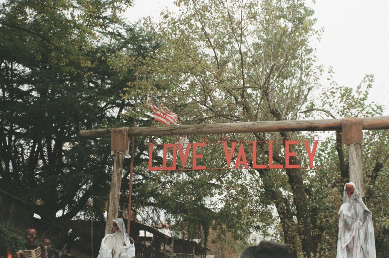 Decorative outdoor entrance with a sign reading 'LOVE VALLEY' hanging on a wooden frame, surrounded by skeleton and ghost Halloween decorations, trees, and an American flag in the background.