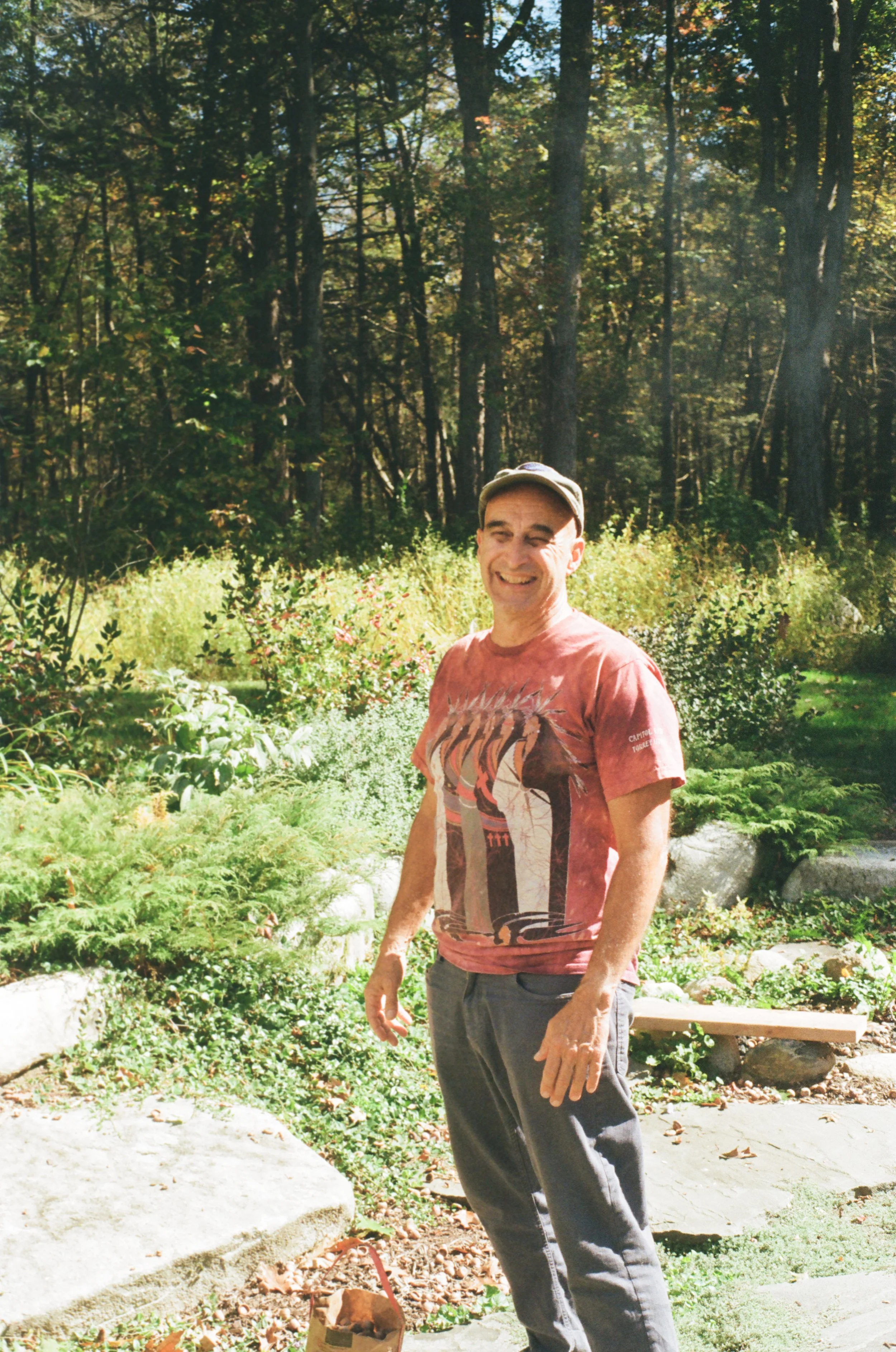 A man smiling outdoors in a garden with green plants and trees in the background.