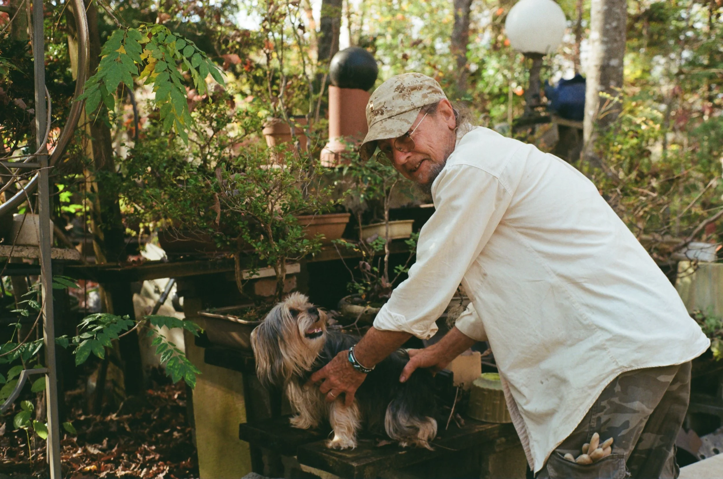 An elderly man with glasses, wearing a camouflage cap and a light-colored shirt, interacts with a small, long-haired dog on a wooden platform in a garden with plants and trees in the background.