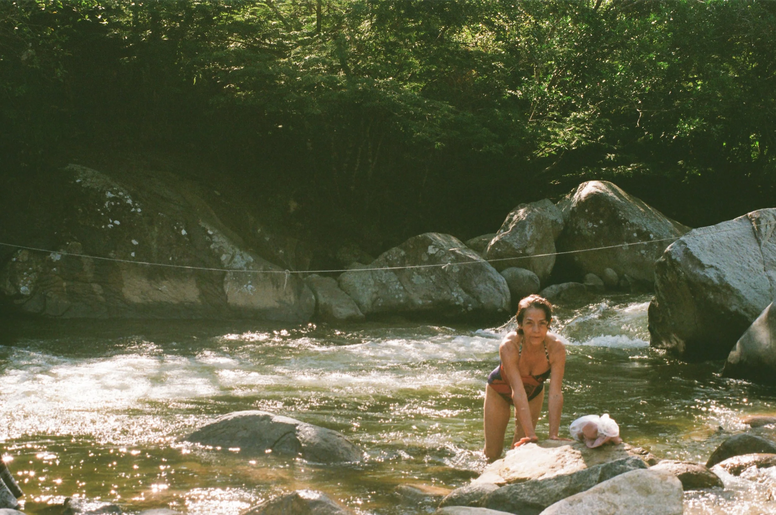 Woman in a swimsuit standing in a rocky river surrounded by trees with sunlight filtering through the leaves.