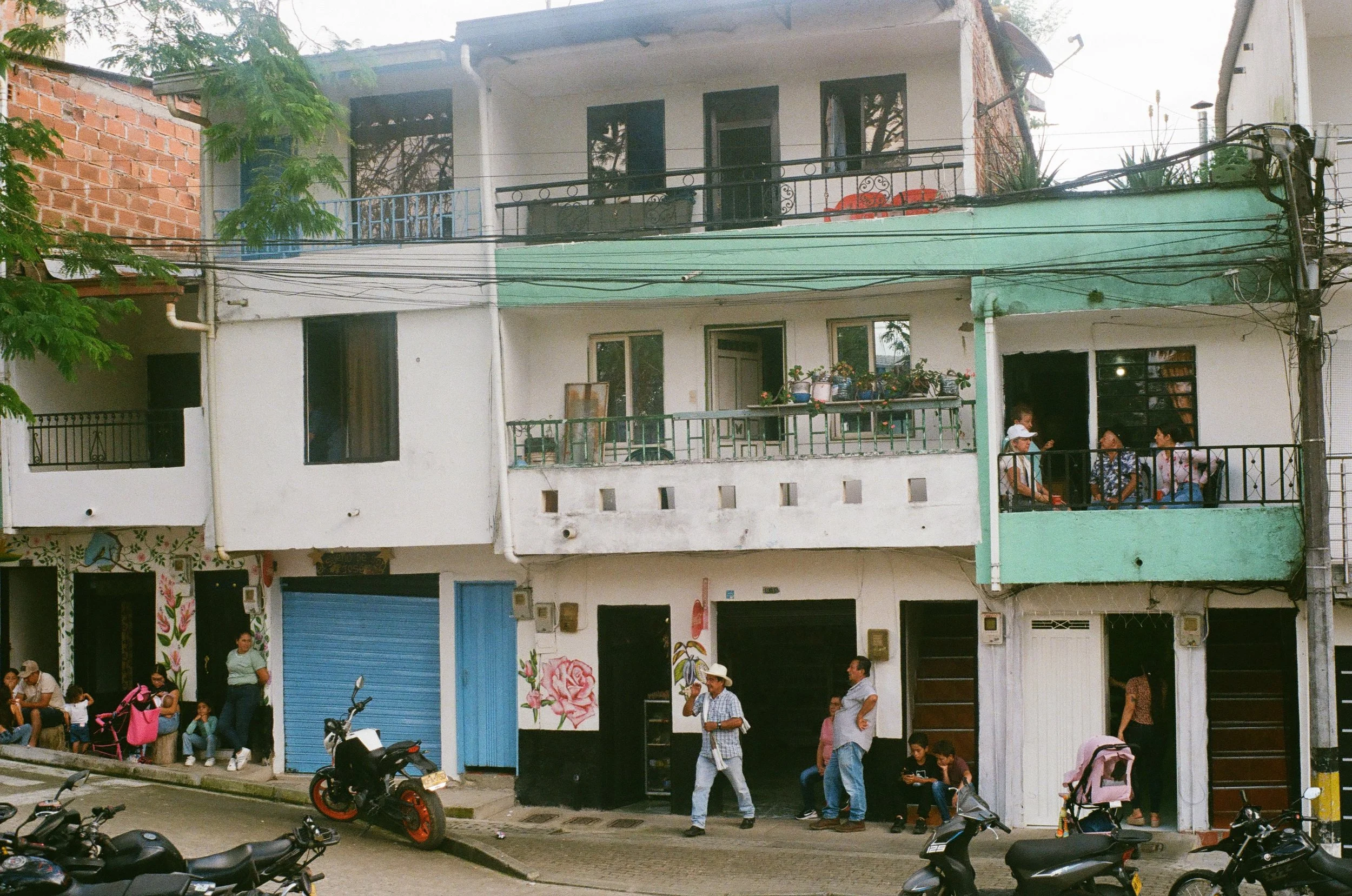 Street scene with two-story buildings, people sitting on the sidewalk, and motorcycles parked along the curb.