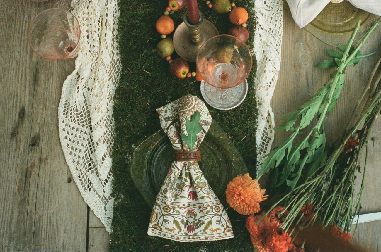 A rustic dining table with a lace table runner, pink wine glasses, a silver candle holder with a red candle, decorative apples, a cloth napkin with a leaf, a clear glass plate, and a bunch of orange and red flowers on the side.